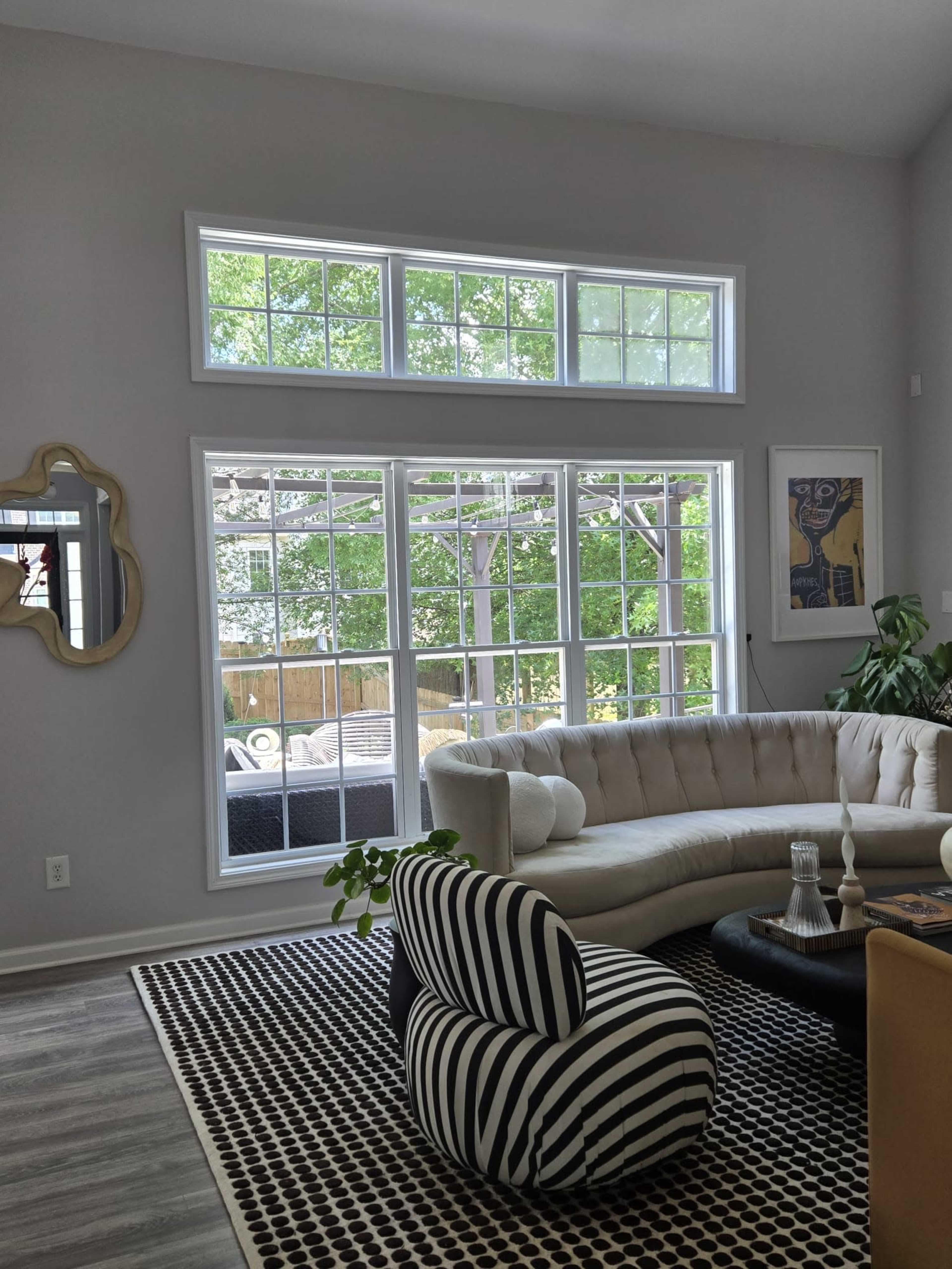 The image shows a modern living room with large windows allowing natural light, featuring a curved sofa, a striped accent chair, and a decorative rug.