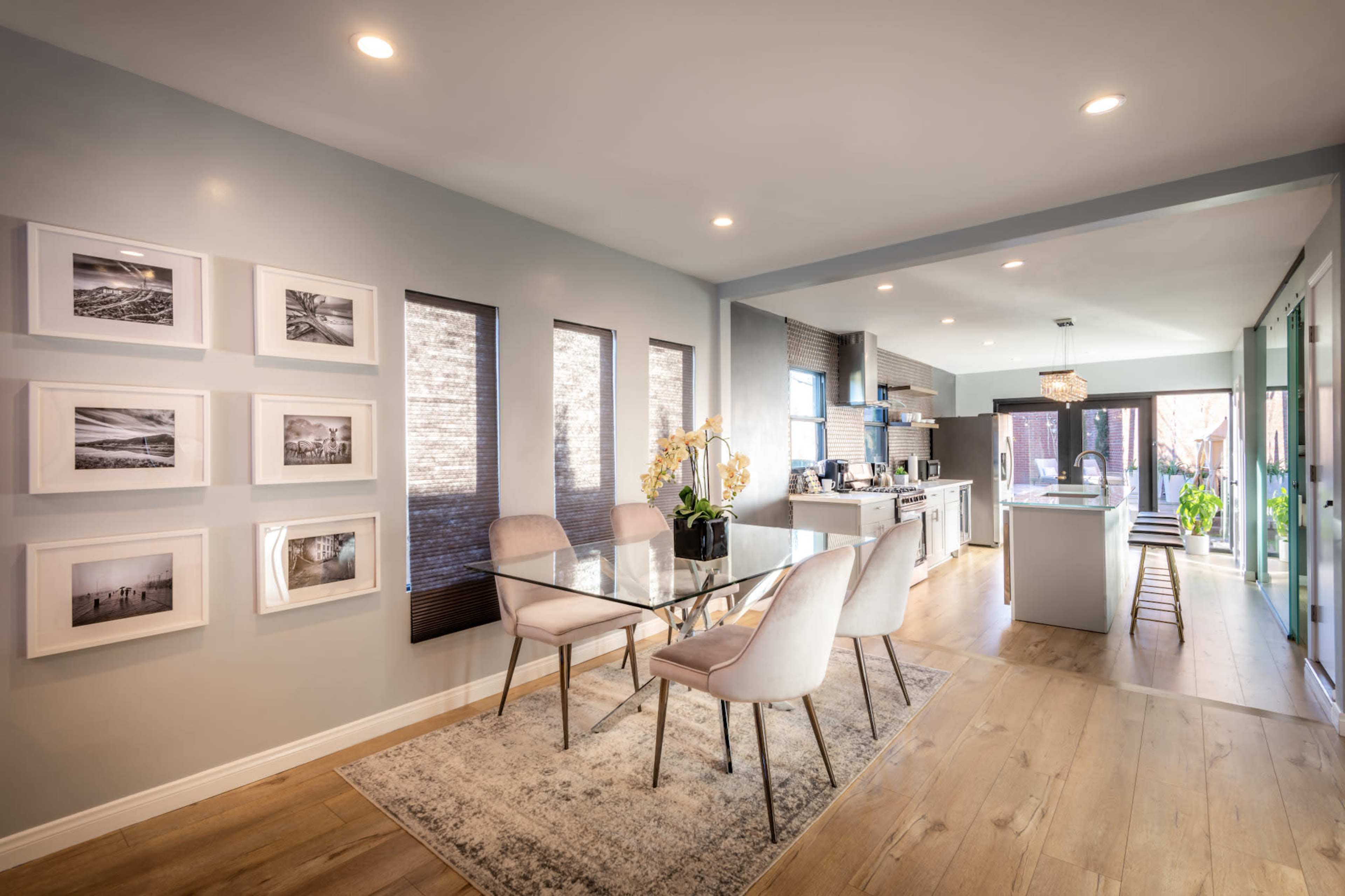 The image shows a modern dining area featuring a glass table surrounded by four chairs, with framed photographs on the wall and an open kitchen in the background.