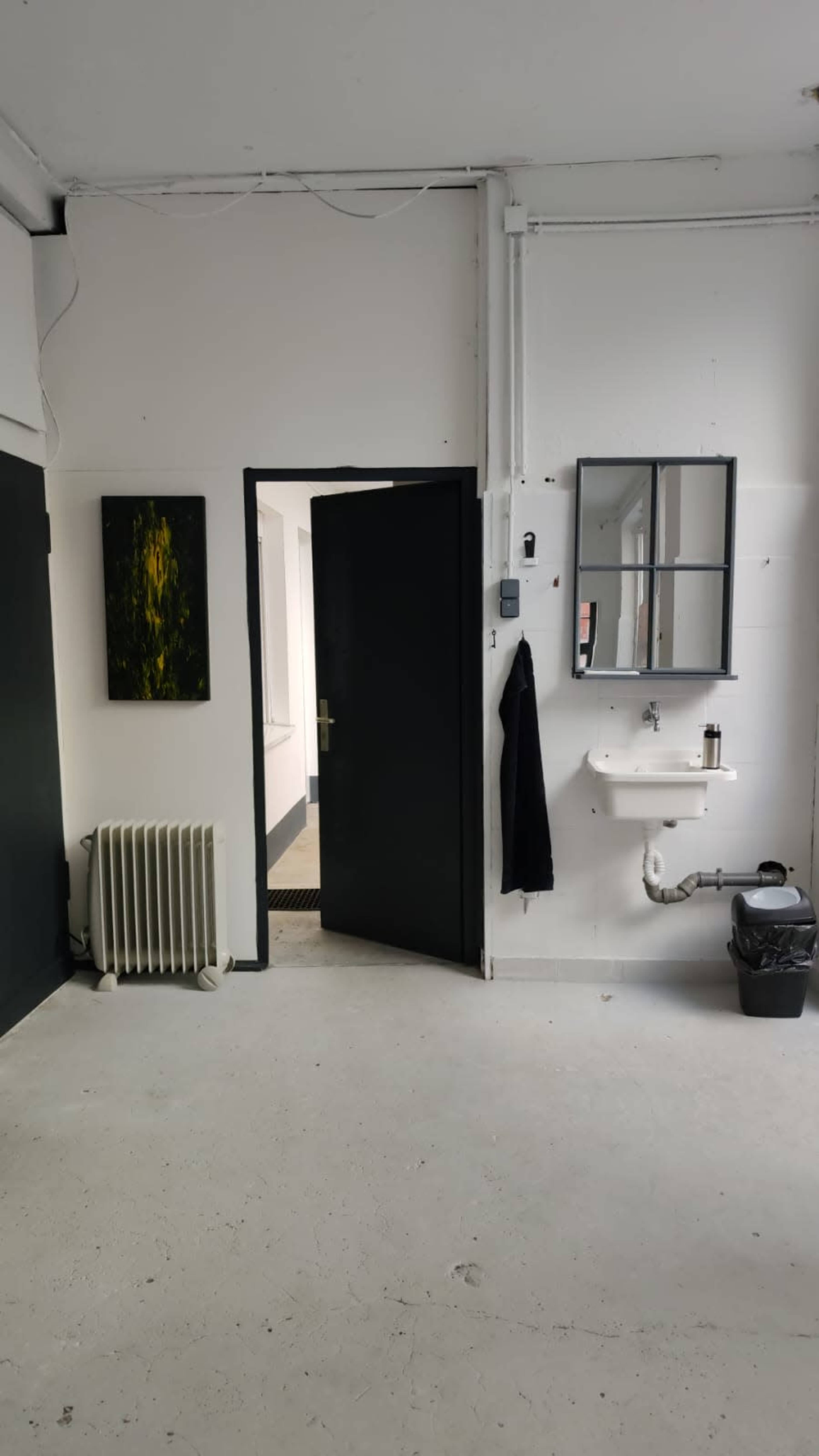 The image shows a minimalistic room with a black door, a white wall featuring a painting, a radiator, a small sink with a mirror, and a trash bin.
