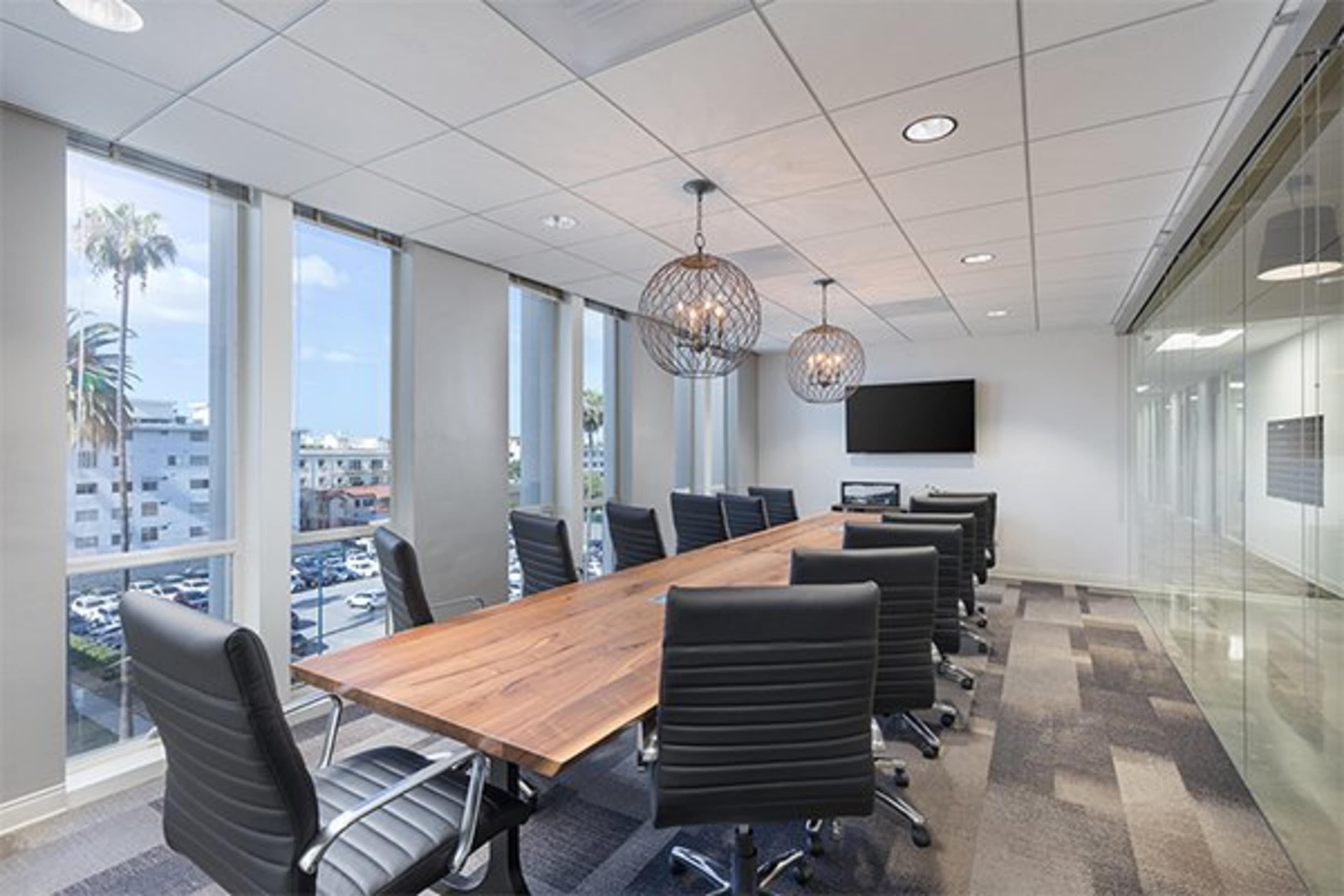 A modern conference room features a large wooden table surrounded by black chairs, with windows offering a view of palm trees and a parking lot outside.