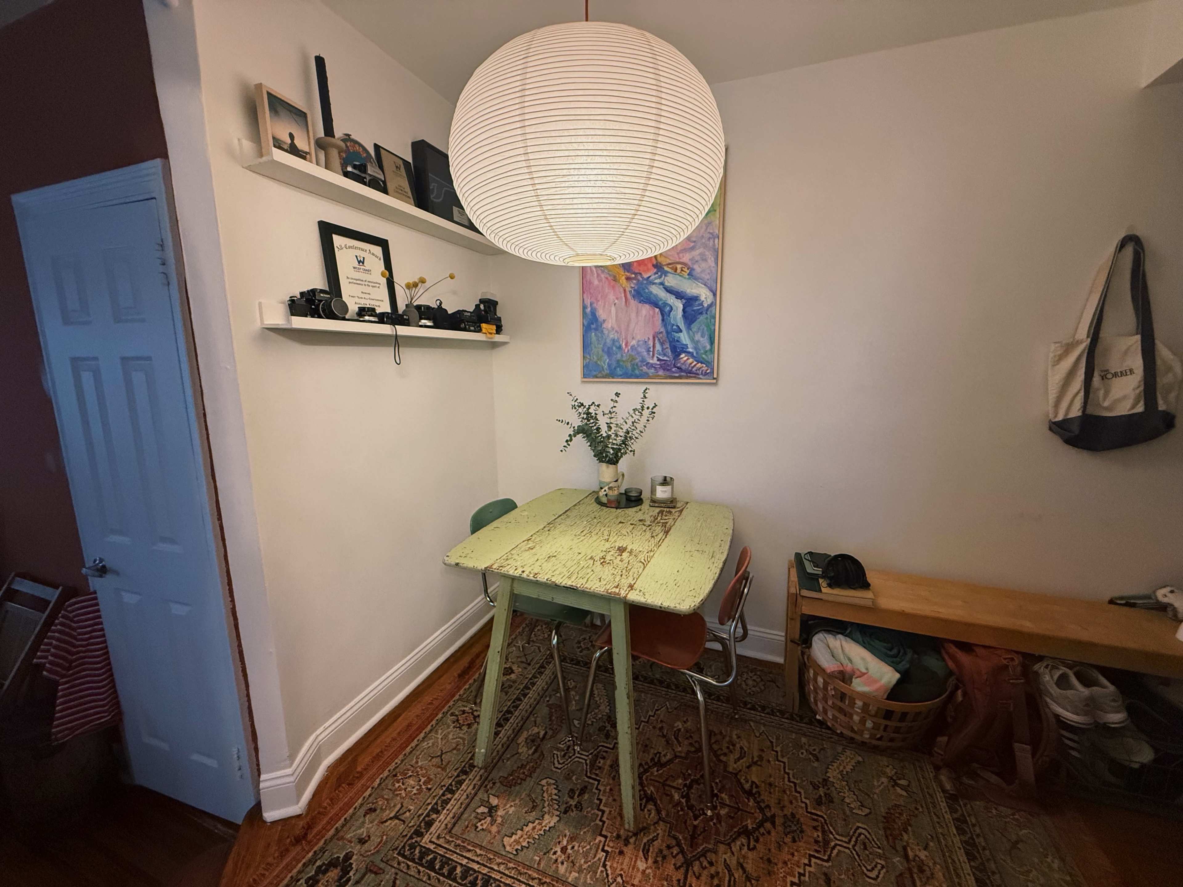 A small dining area features a green wooden table with two chairs, a pendant light above, and a shelf displaying various items on the wall.