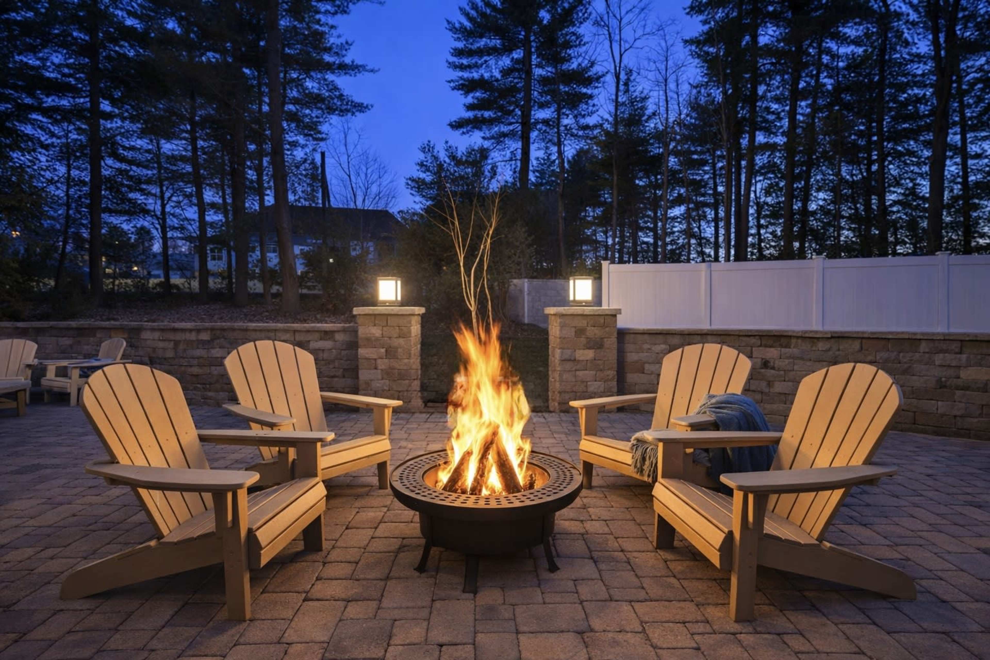 A circular fire pit surrounded by four wooden chairs is illuminated by a fire at dusk, with trees in the background.