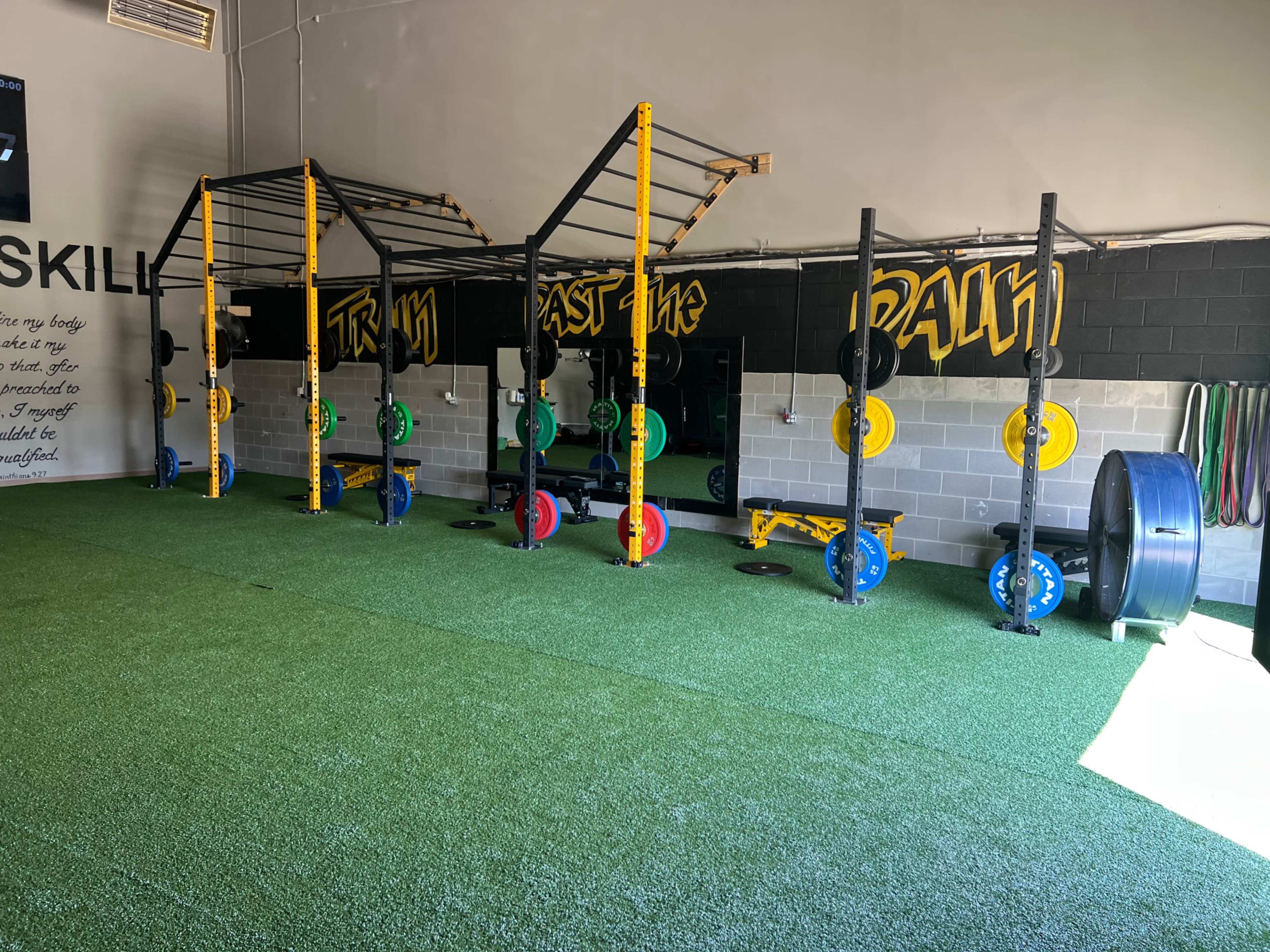 A gym interior featuring weightlifting equipment, including squat racks with colorful weight plates, a pull-up bar structure, and artificial turf flooring.