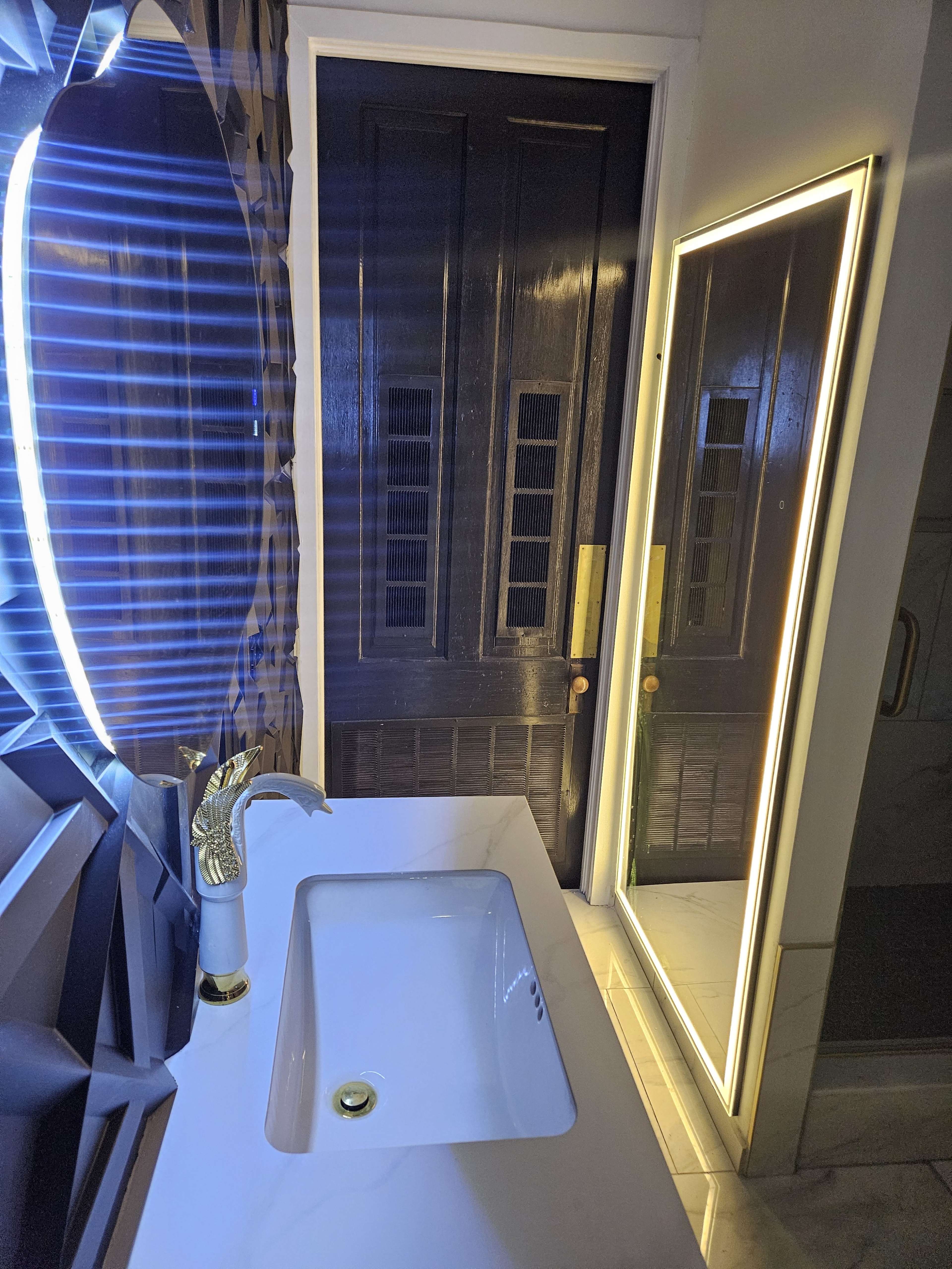 The image shows a modern bathroom sink with a sleek faucet, a tall mirror with LED lighting, and a dark wooden door in the background.