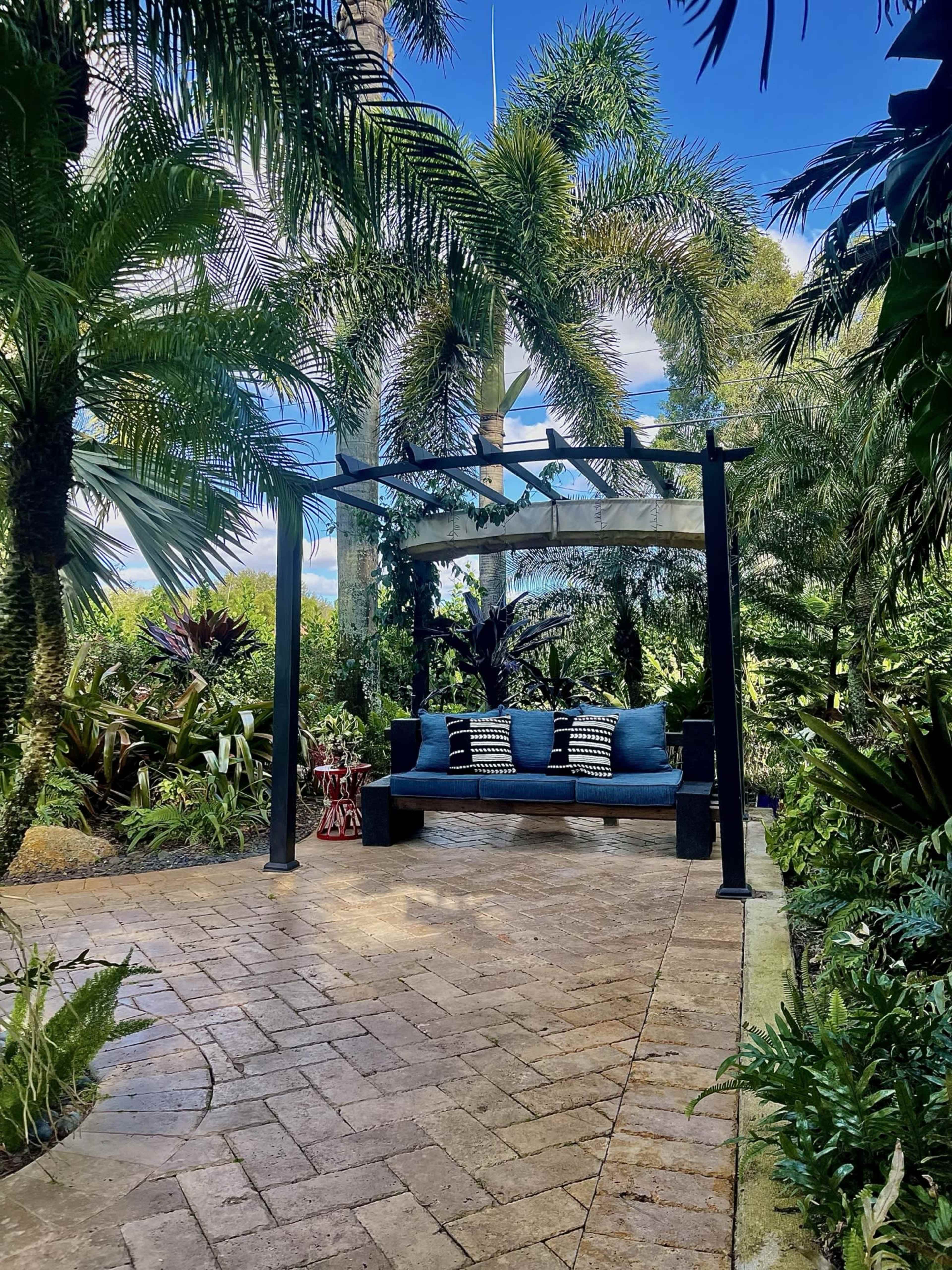 A sheltered seating area with a blue couch and decorative pillows is surrounded by lush tropical plants and palm trees.