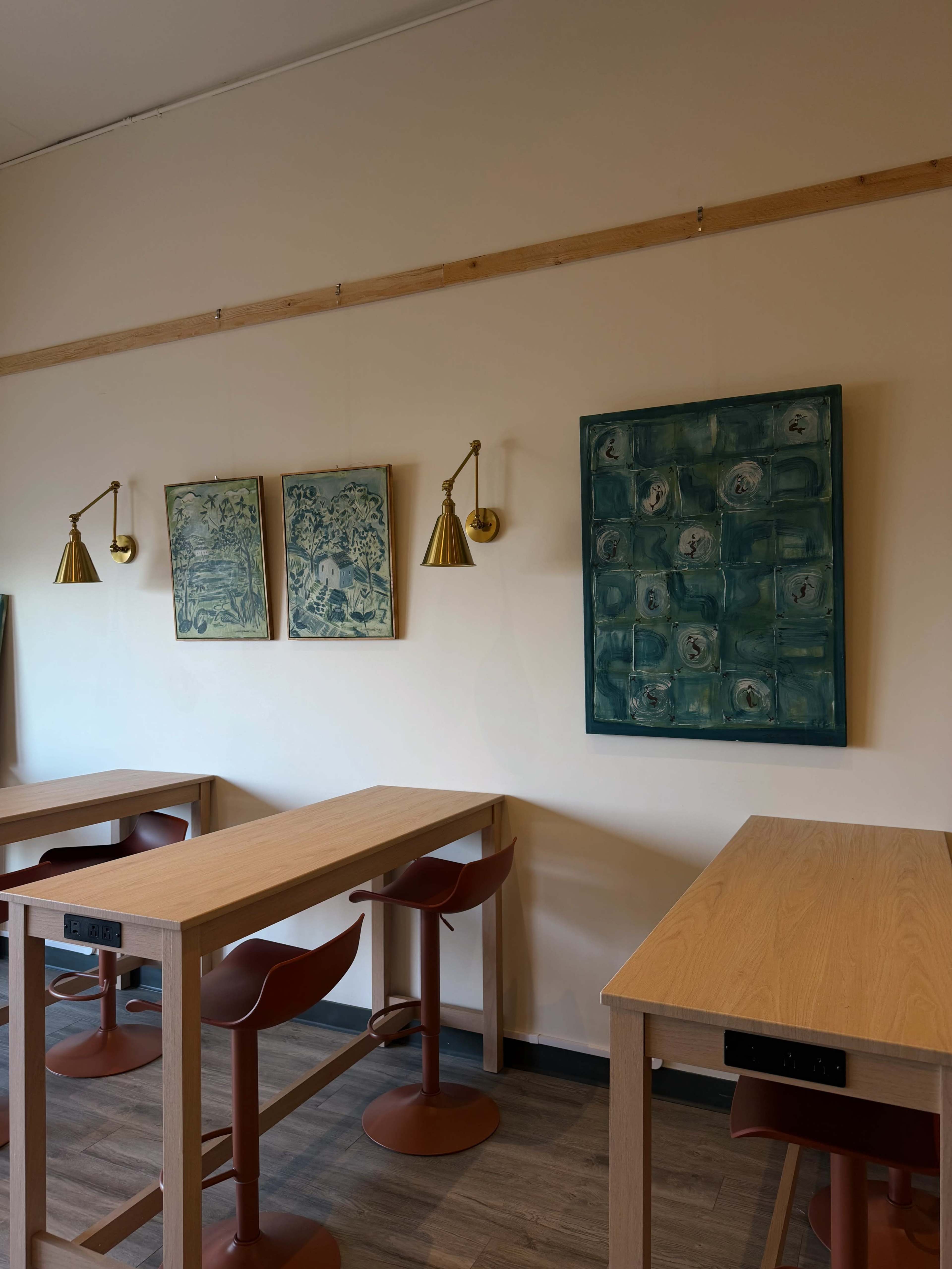 The image shows a cozy café interior with wooden tables, red chairs, and wall-mounted lamps, featuring three pieces of framed artwork on the walls.