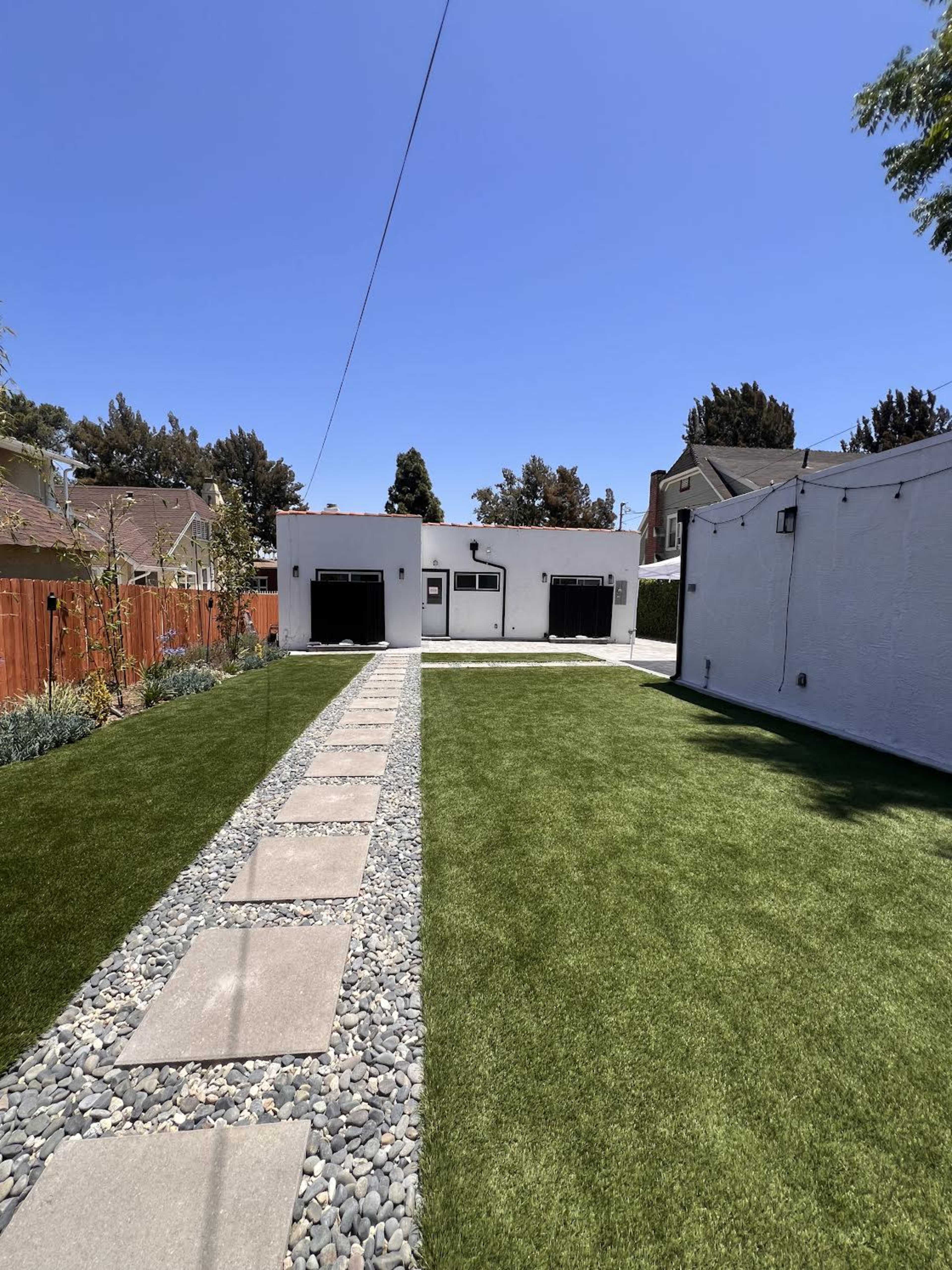 The image shows a well-maintained backyard featuring a path made of stepping stones lined with gravel, leading to a white building under a clear blue sky.