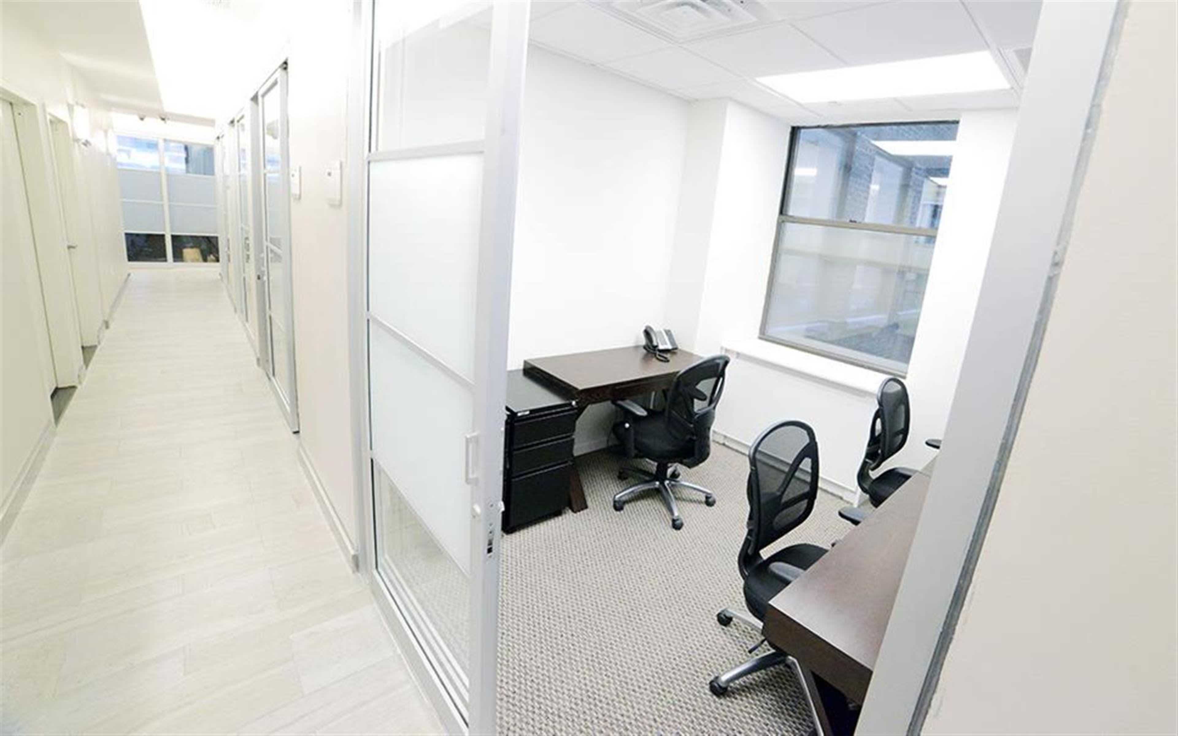 A bright office space with two desks and chairs arranged in a room, adjacent to a hallway lined with doors.