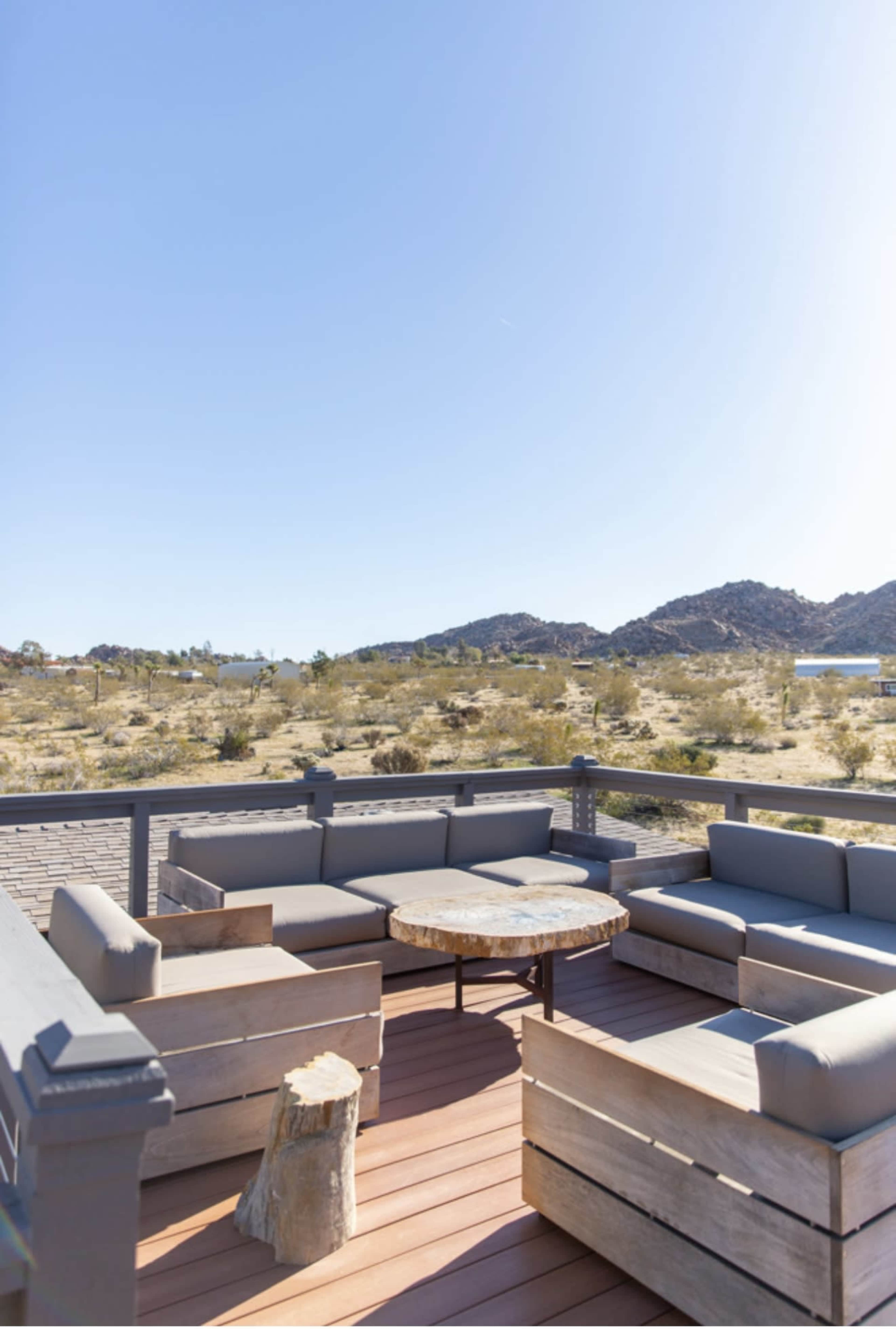 A wooden patio area features gray cushioned seating arranged around a circular coffee table, with desert landscape and mountains in the background under a clear blue sky.