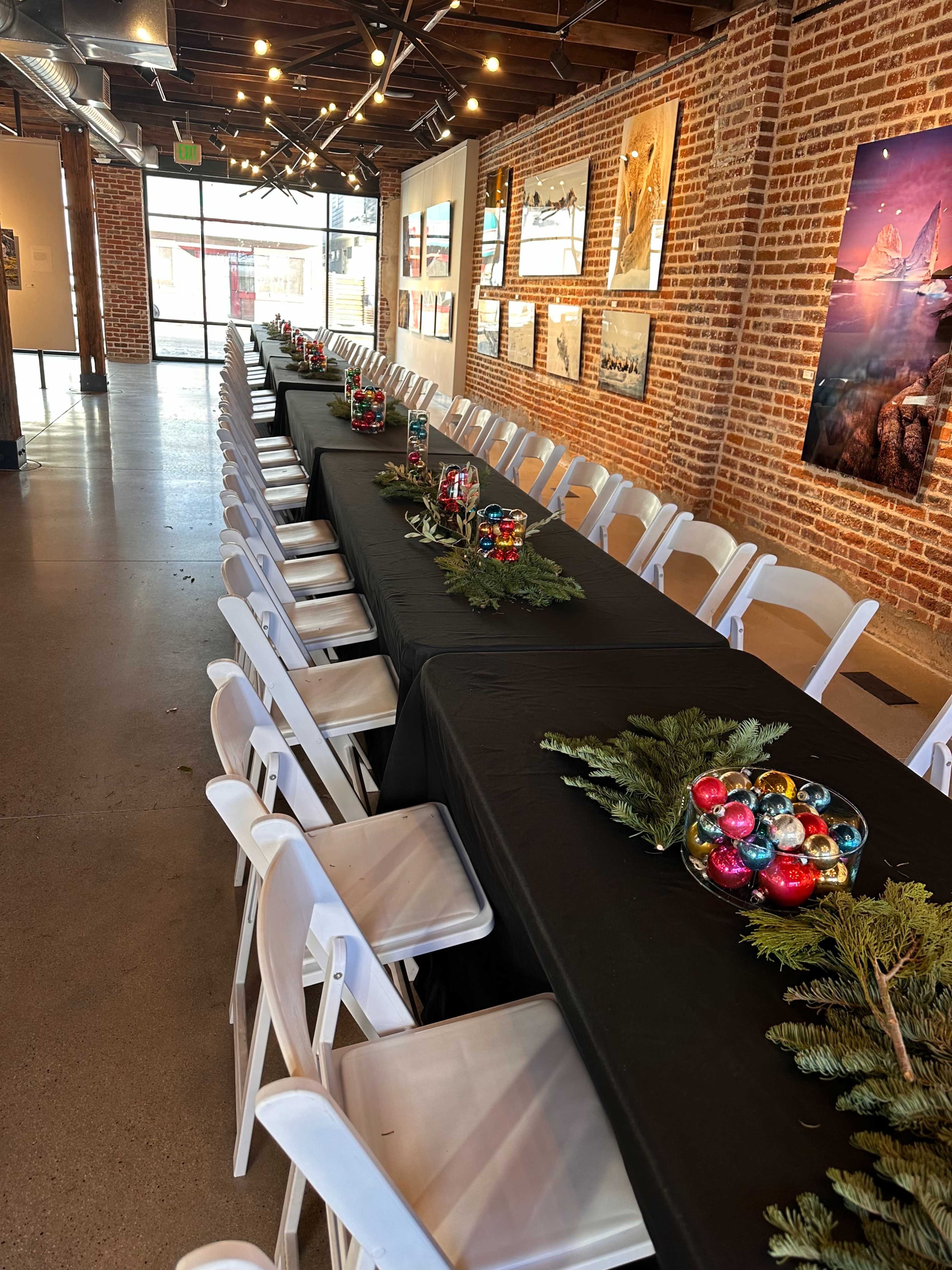 A long table is set with black tablecloths and decorated with colorful ornaments and greenery in a brick-walled event space.