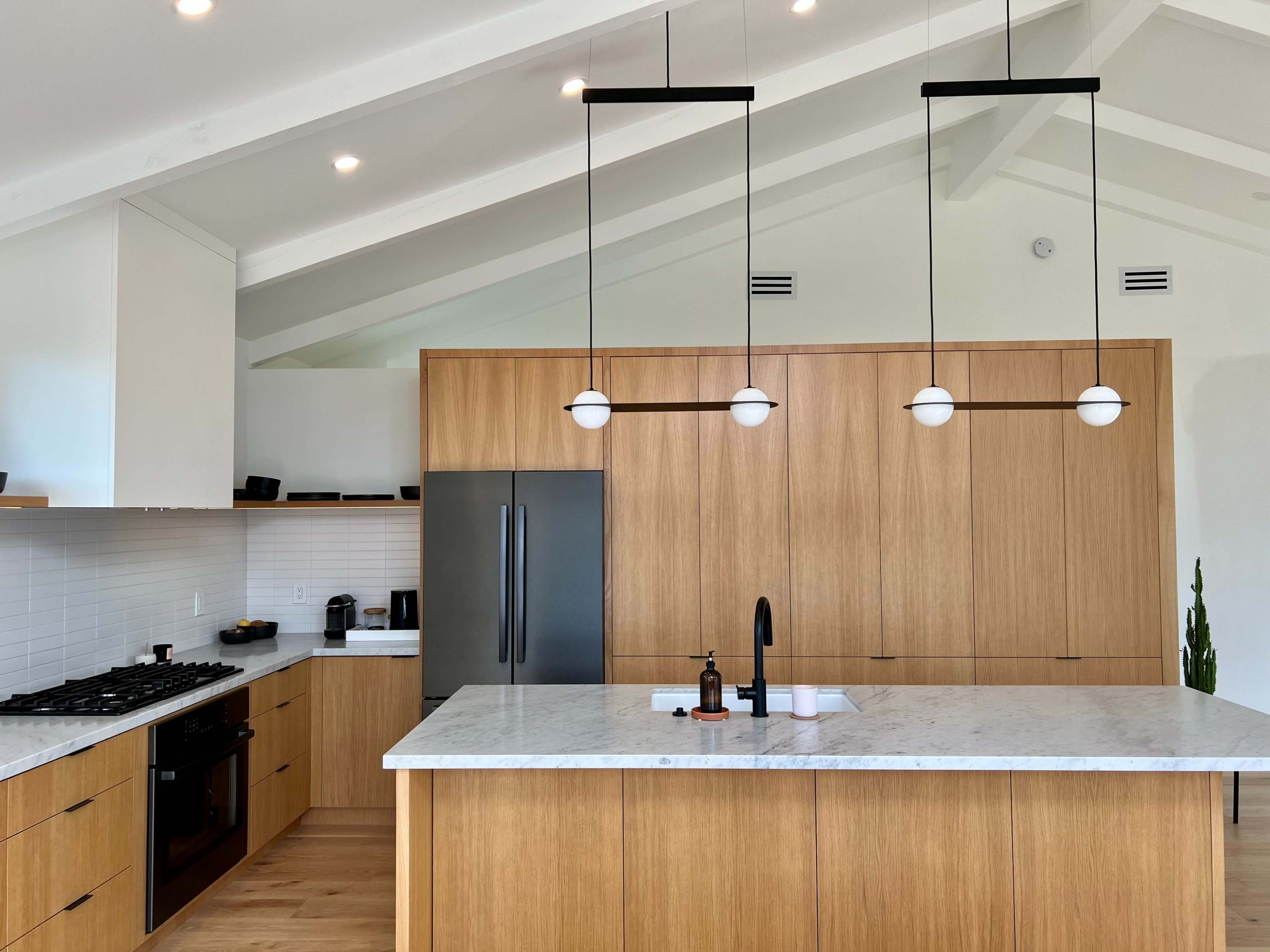 A modern kitchen features an island with a marble countertop, wooden cabinetry, and pendant lights hanging from a vaulted ceiling.