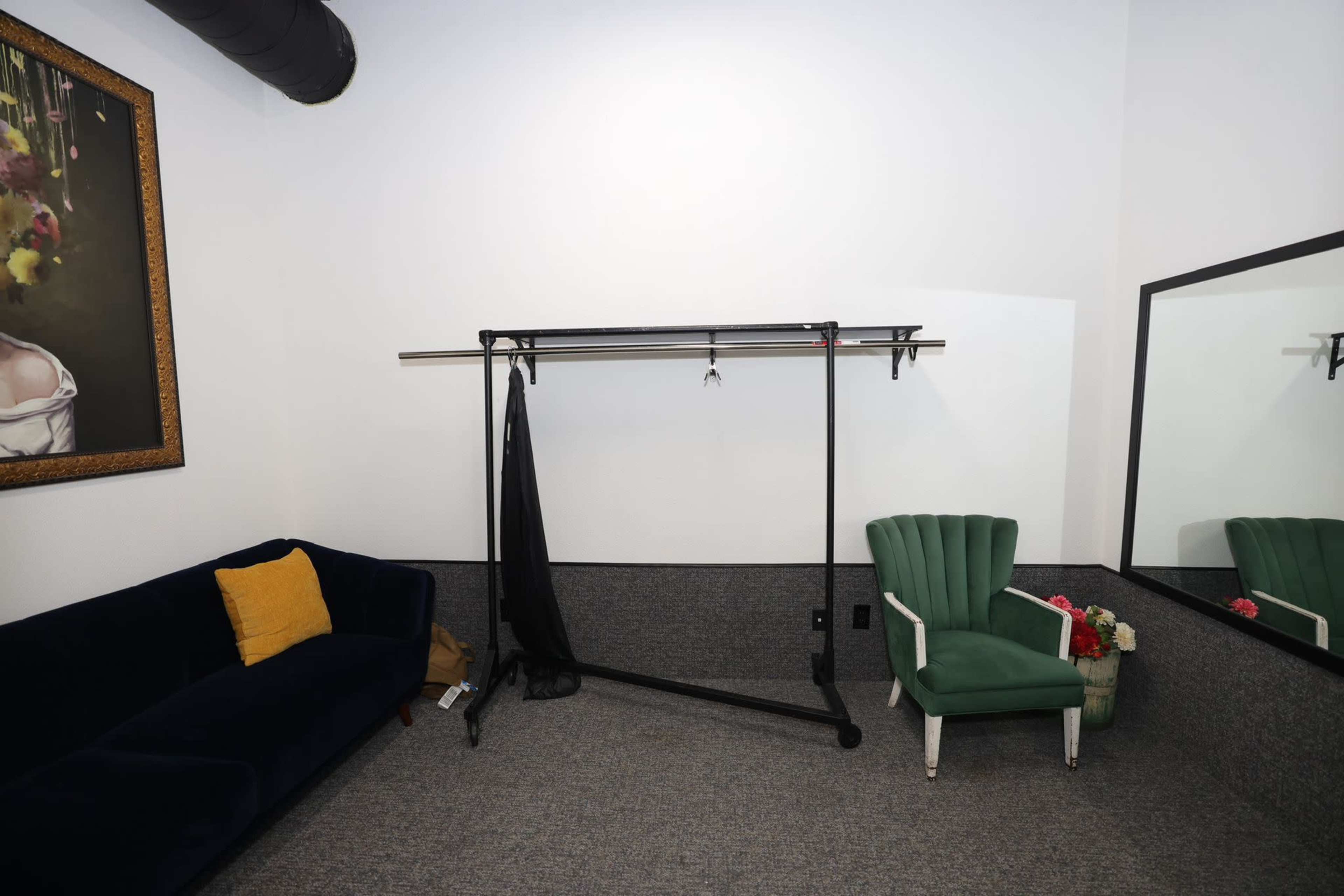 A minimalist room with a dark green couch, a clothing rack, a mirror, and a green chair with white trim.