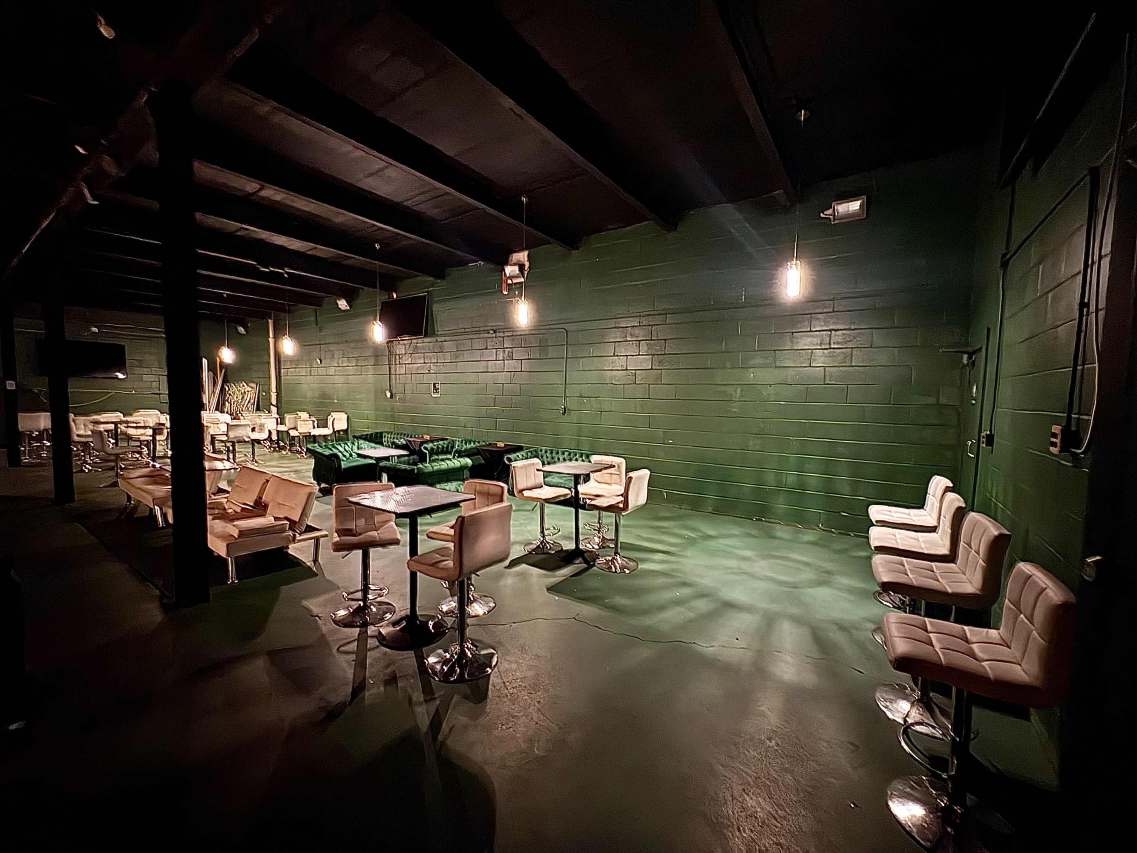 The image shows a dimly lit lounge area with green walls, featuring a mix of seating options including chairs and bar stools arranged around small tables.