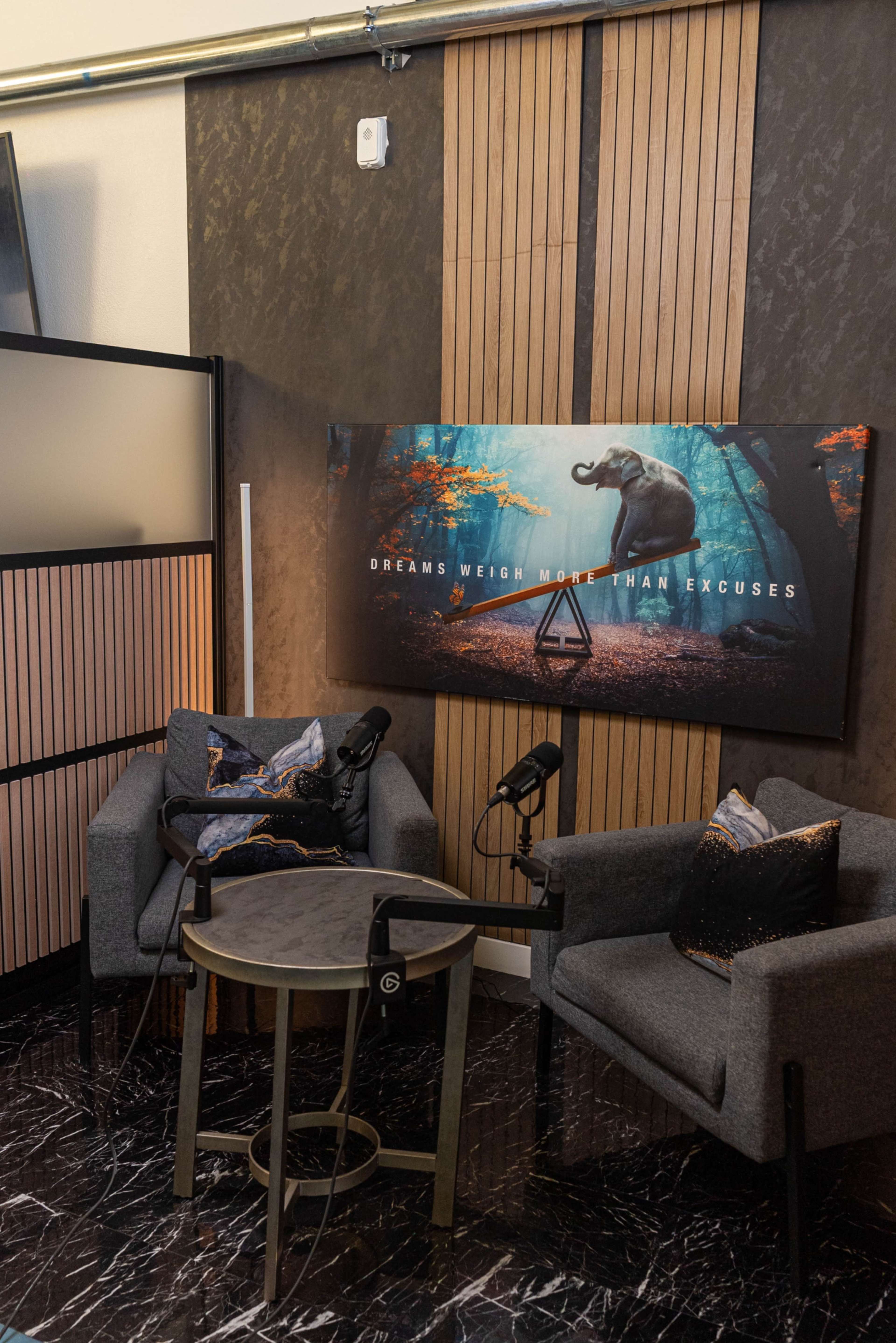 The image shows a cozy interview setup featuring two gray armchairs, a small round table, and a wall-mounted decorative piece with the text "DREAMS WEIGH MORE THAN EXCUSES."