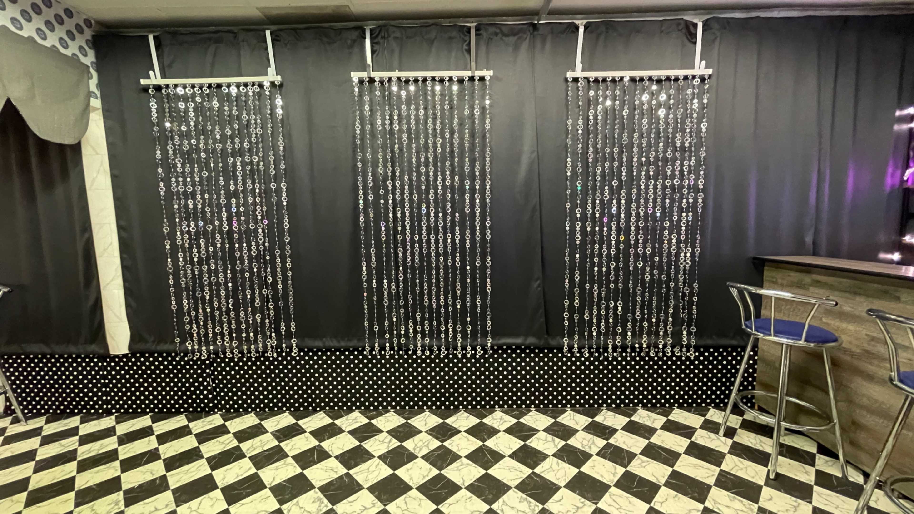 The image shows a room with three vertical strands of hanging beads against a black curtain, with a checkered floor and a bar in the background.