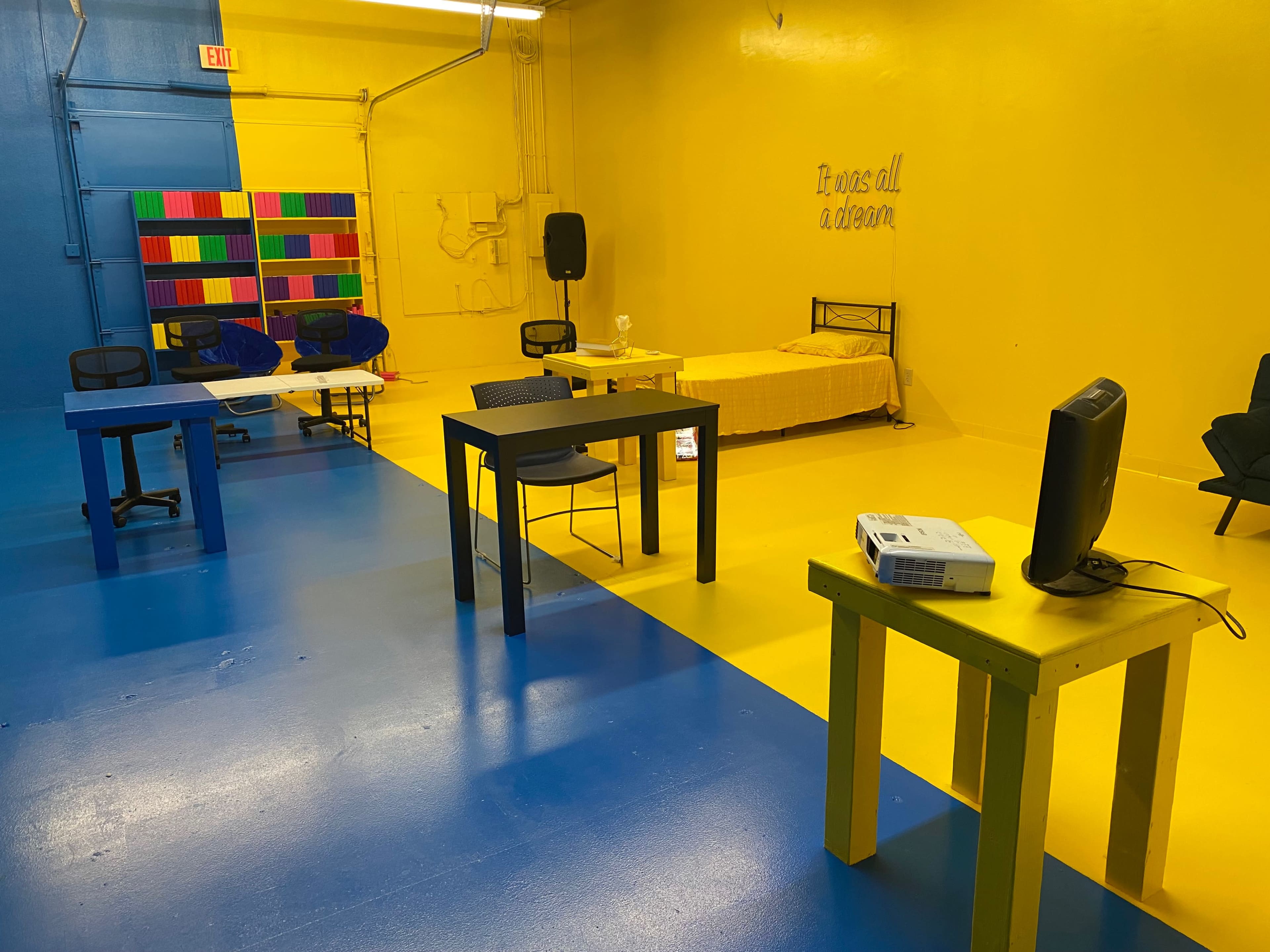 The image shows a brightly colored room with yellow and blue flooring, featuring several tables, chairs, a bed, and a small projector set up.