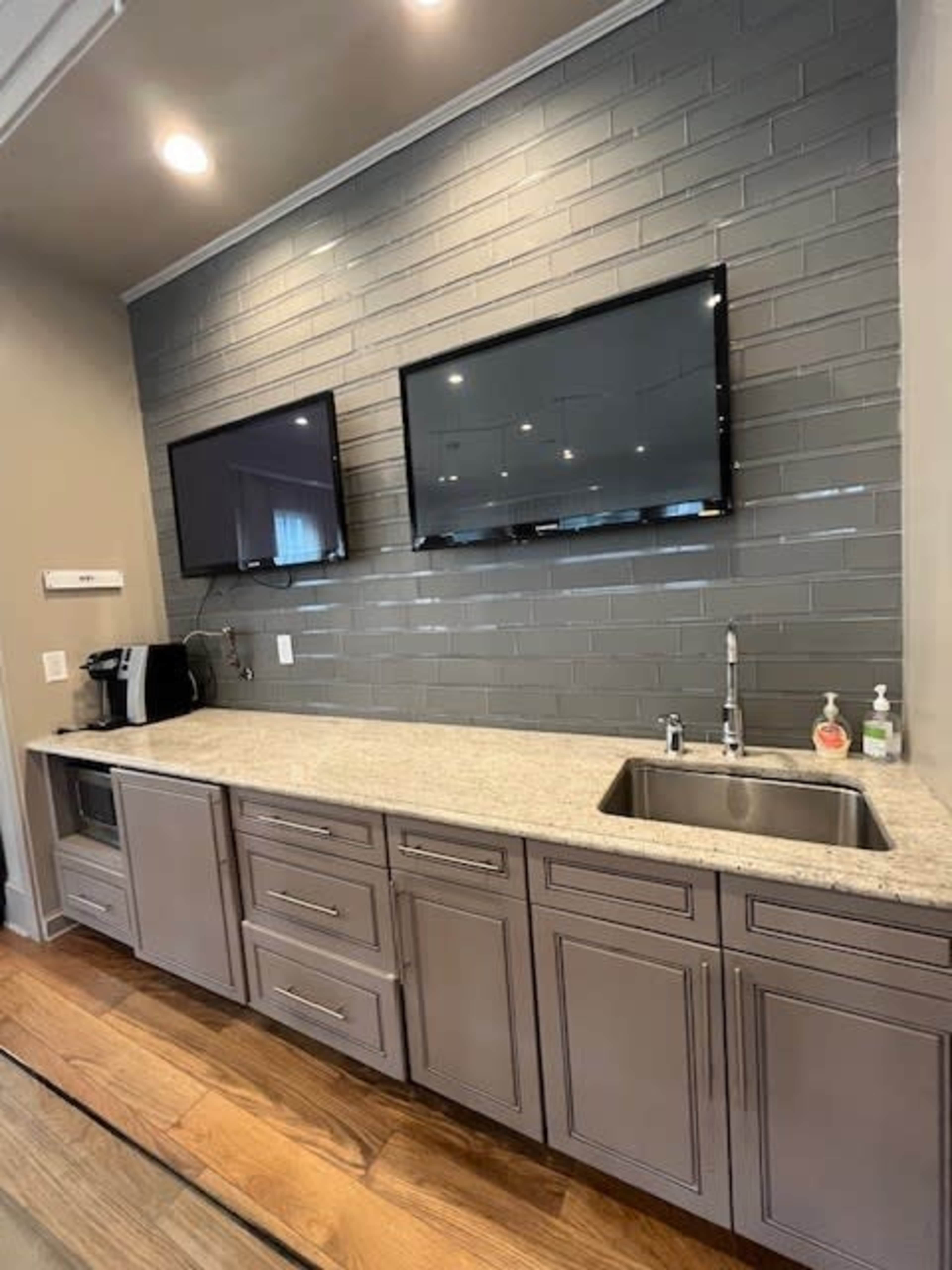 A modern kitchen area with gray tile walls, a granite countertop, two mounted flat-screen TVs, and a sink with a modern faucet.