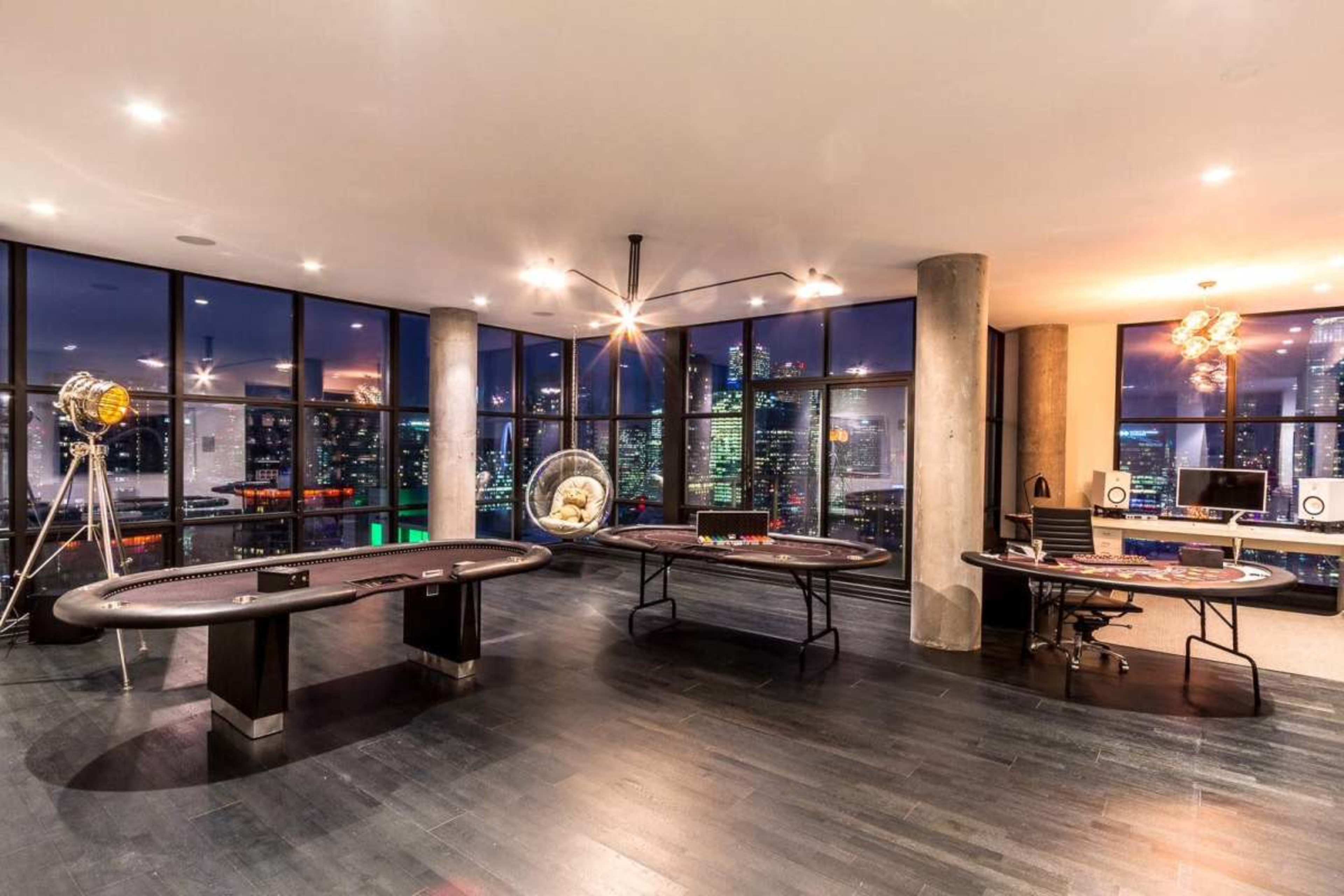 The image shows a sleek game room with poker and blackjack tables, surrounded by large windows displaying a city skyline at dusk.