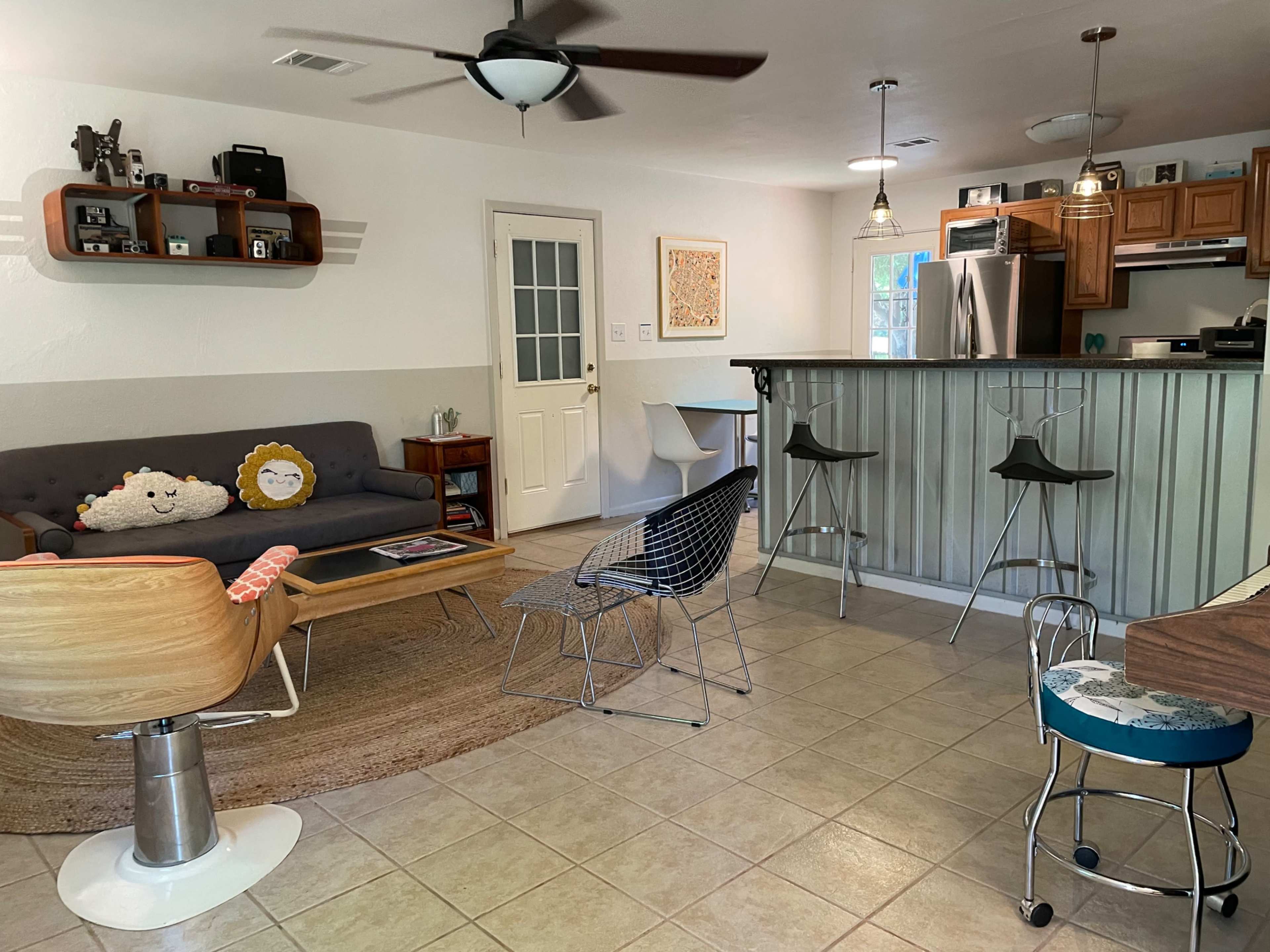 The image shows a modern living area with a sofa, a wooden coffee table, bar stools at a kitchen counter, and a mix of decorative items along with a door leading outside.