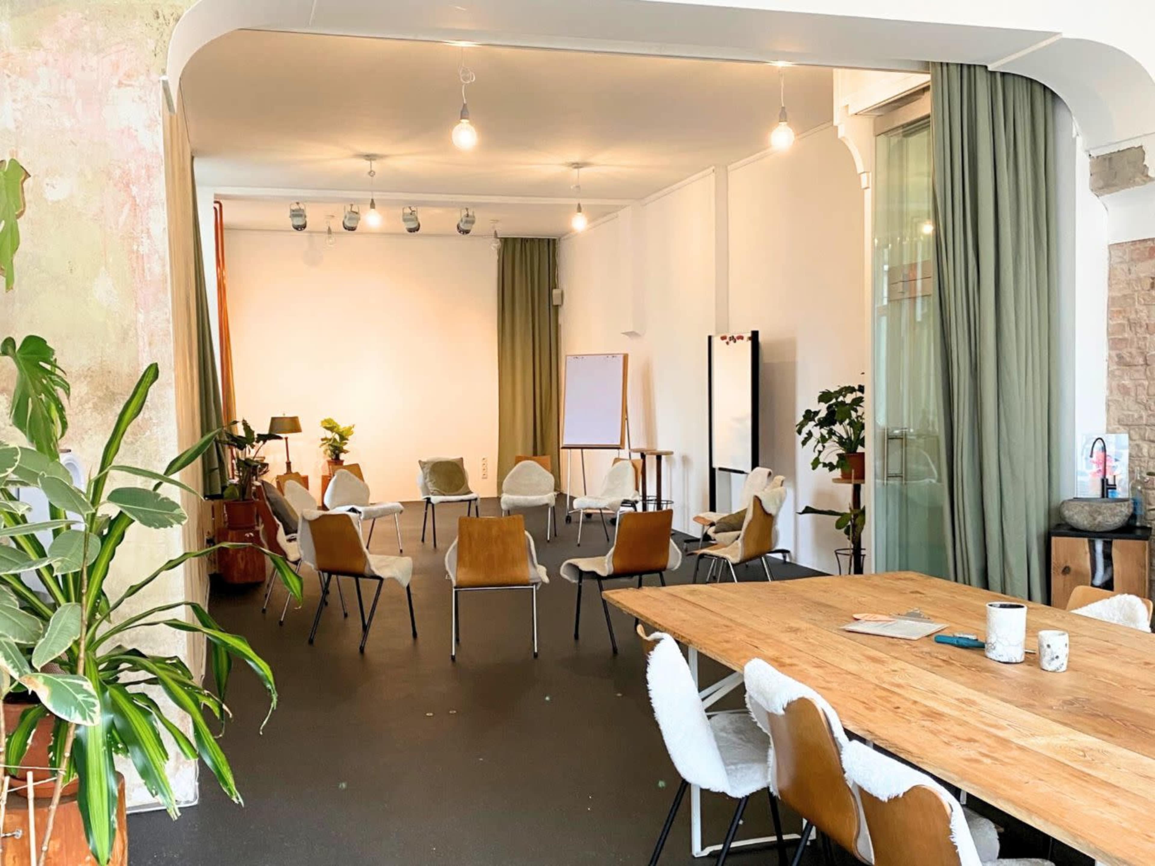 The image shows a spacious meeting room with a wooden table, several chairs arranged in a semicircle, plants, and soft lighting.