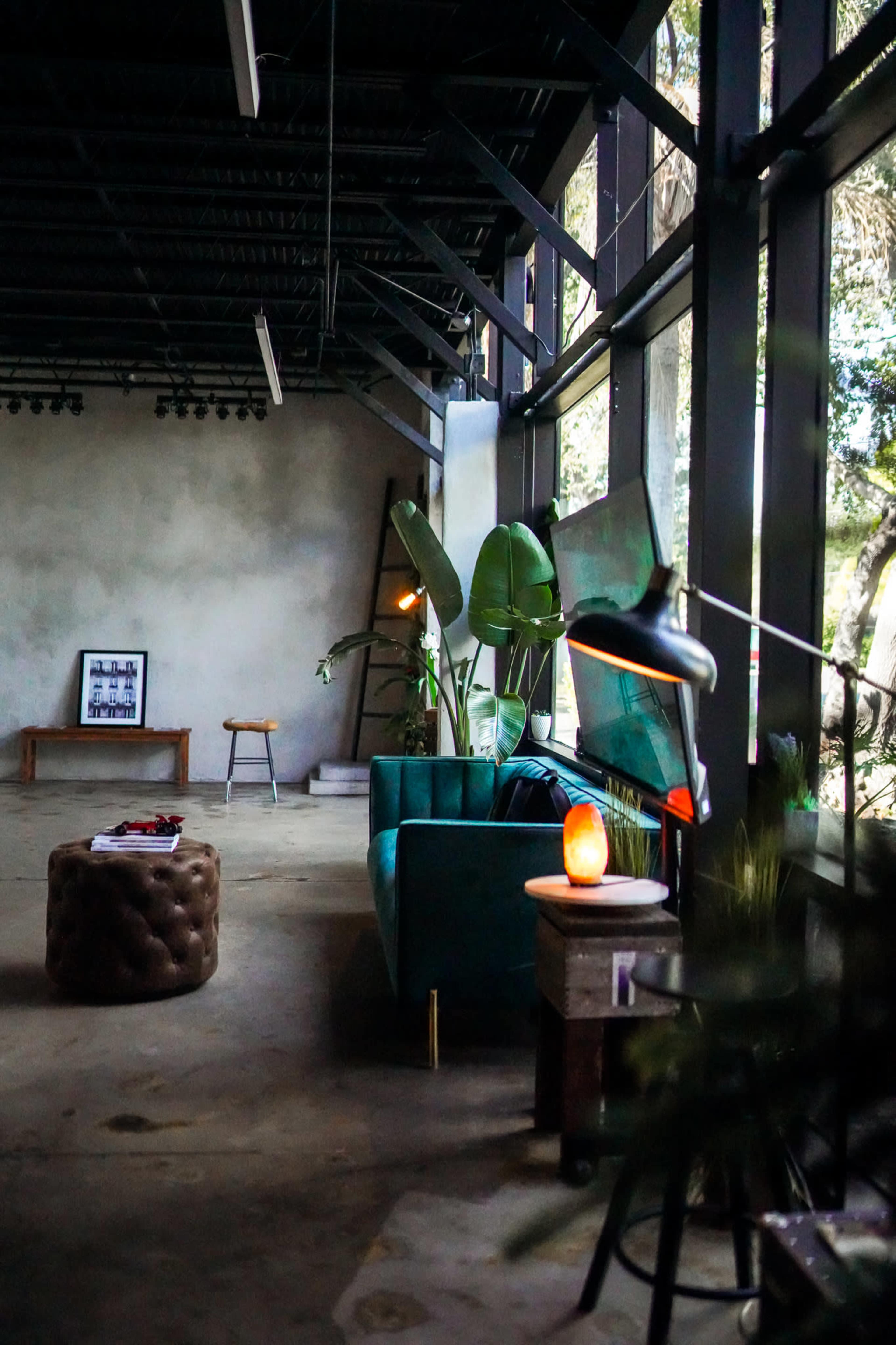 The image shows a spacious room with large windows, featuring a green sofa, a round ottoman, and potted plants, along with minimal furniture against a concrete wall.