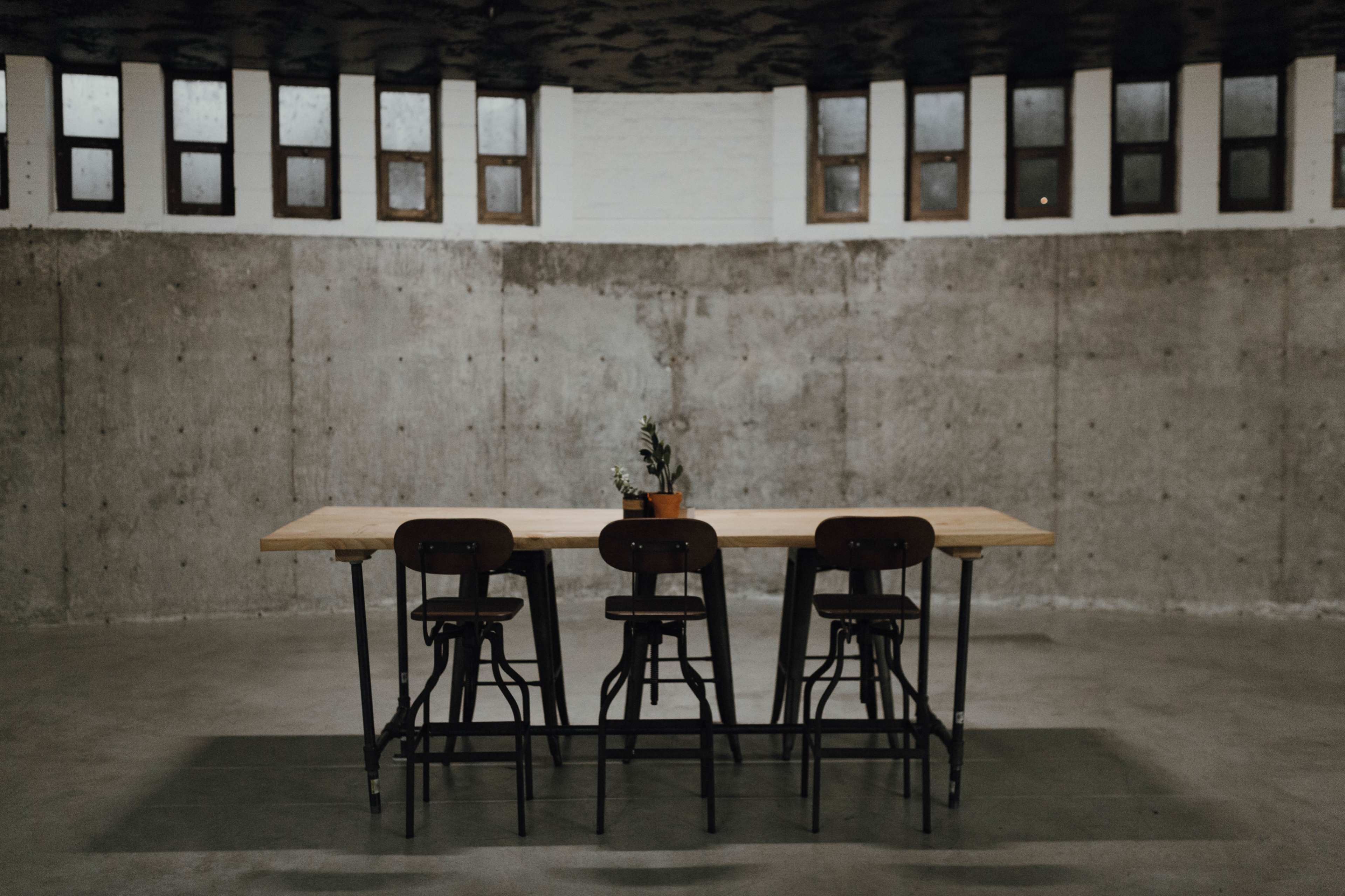 A long wooden table with four chairs is positioned in a room featuring concrete walls and large windows.