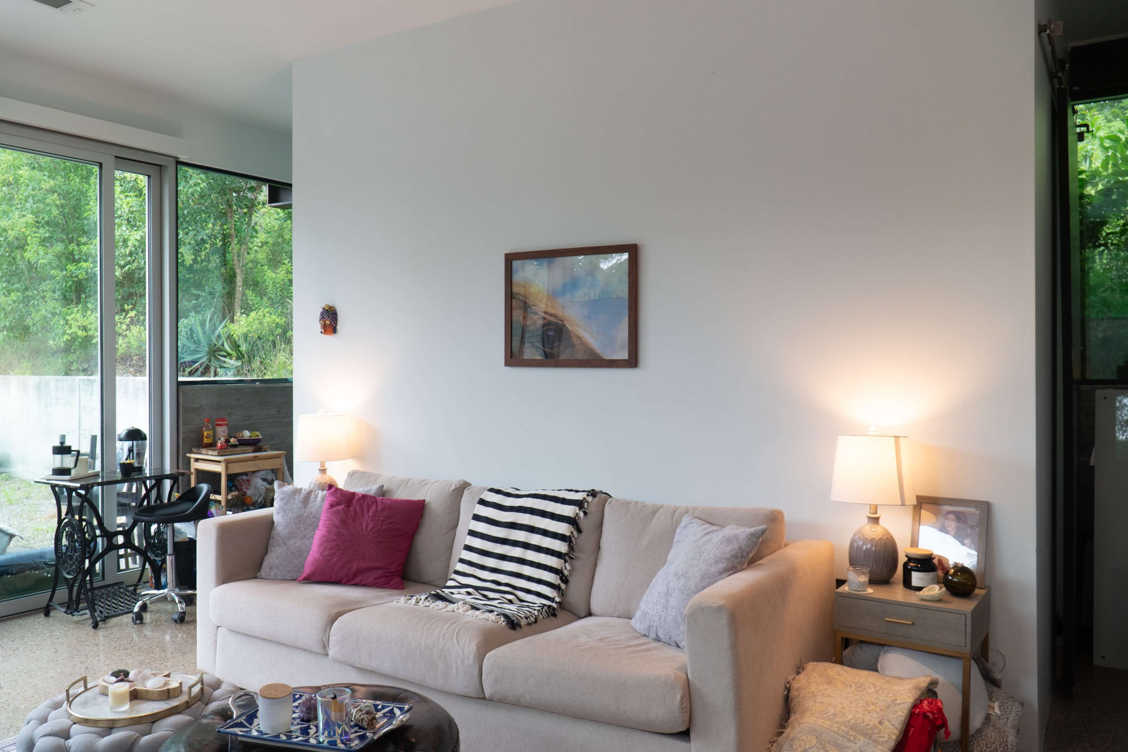 A modern living room features a beige sofa with decorative pillows, a coffee table with items on it, and large windows that overlook a green outdoor area.