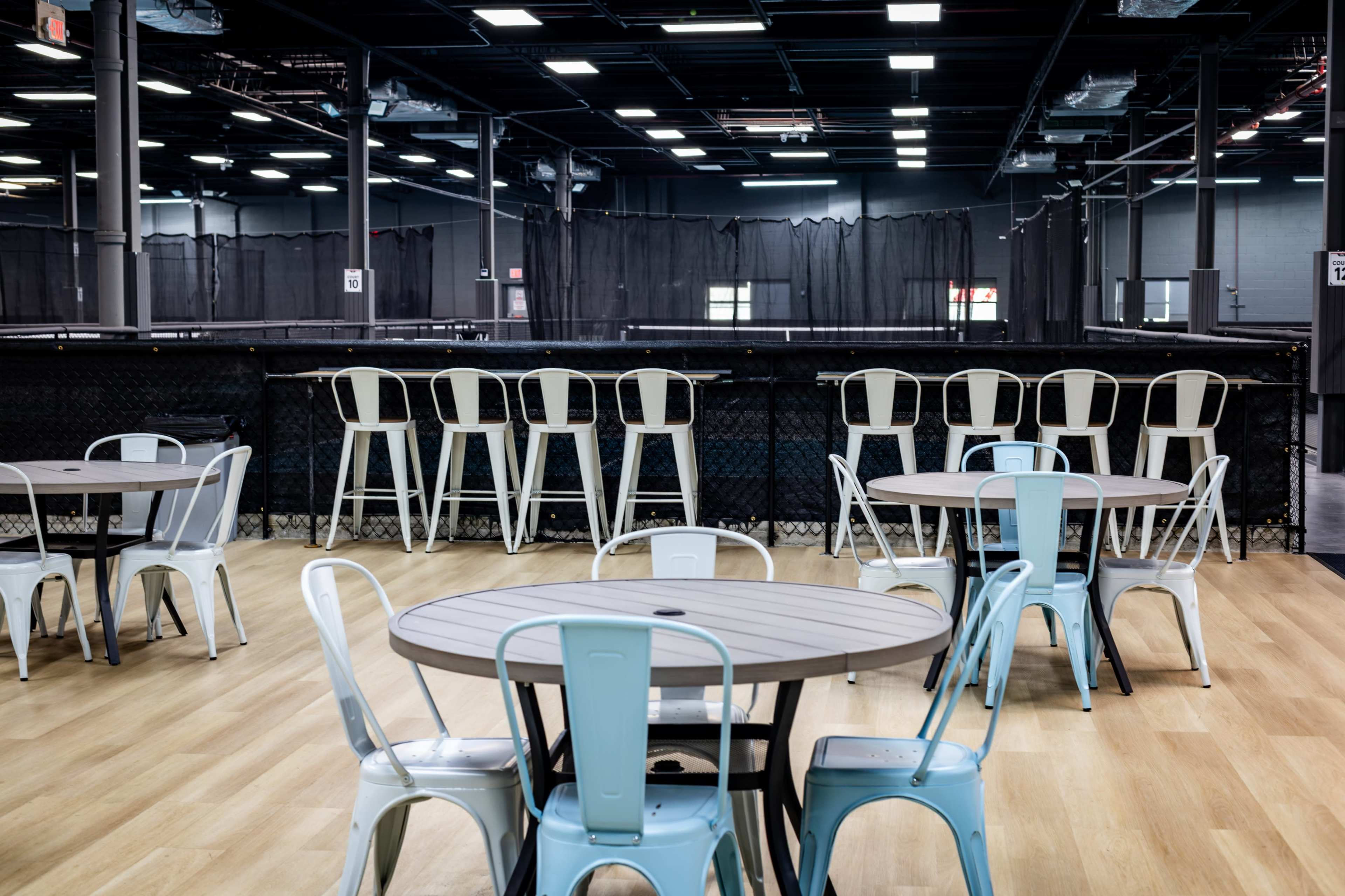 The image shows a spacious indoor venue with several round tables and metal chairs, with a backdrop of a netting area and ample overhead lighting.