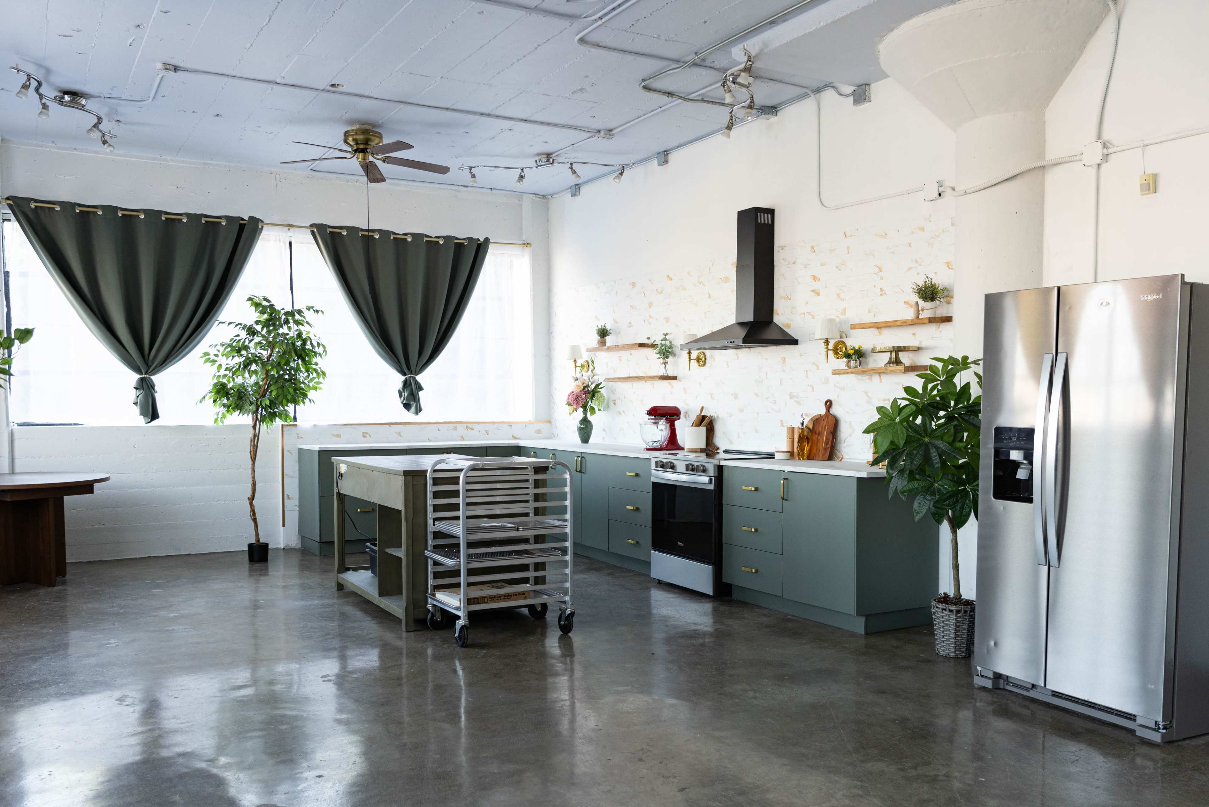 The kitchen features a large central island with an oven, a refrigerator, and green cabinetry, set against a backdrop of large windows and potted plants.