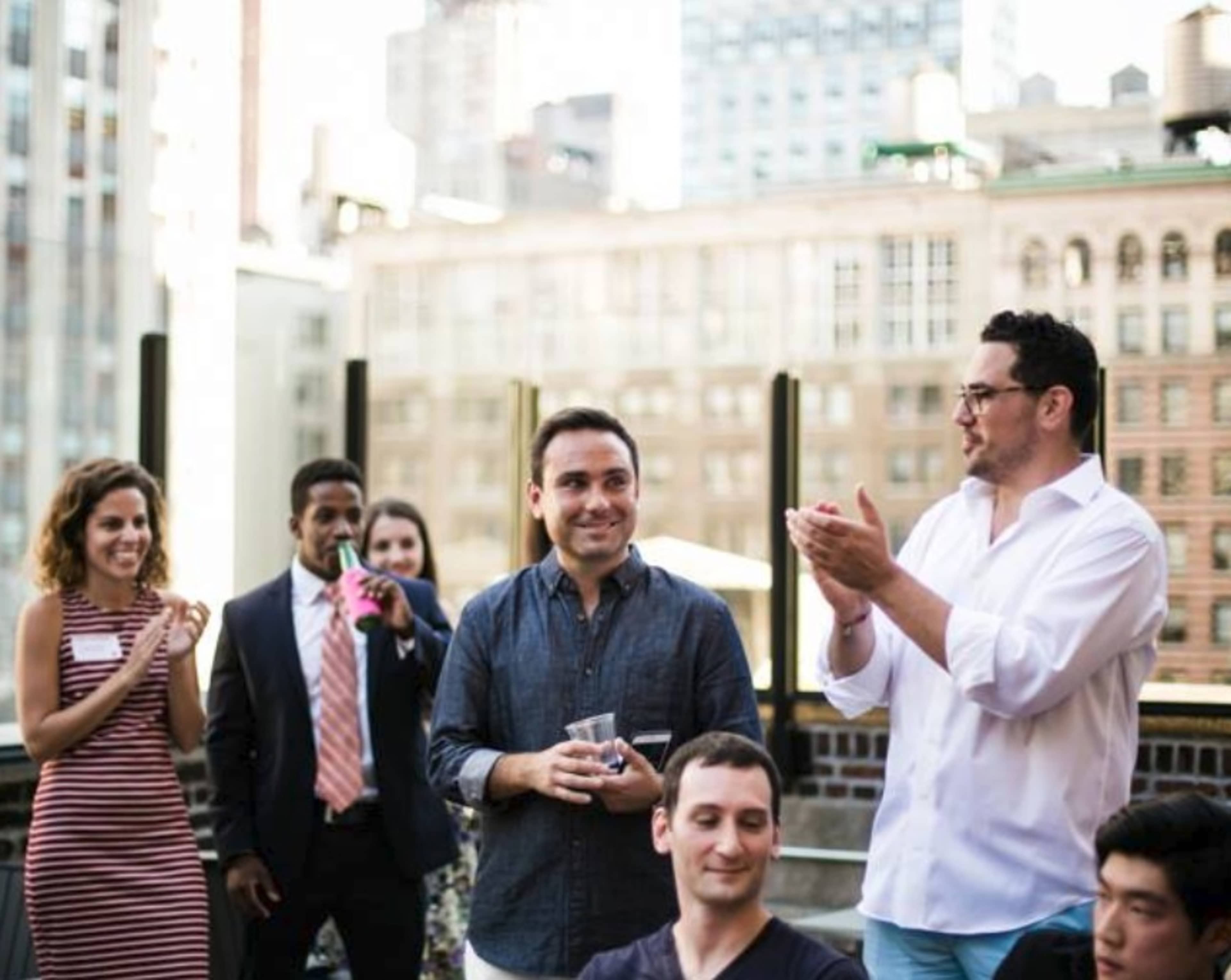 A group of people gathers on a rooftop, with some applauding and others smiling, in an urban setting.