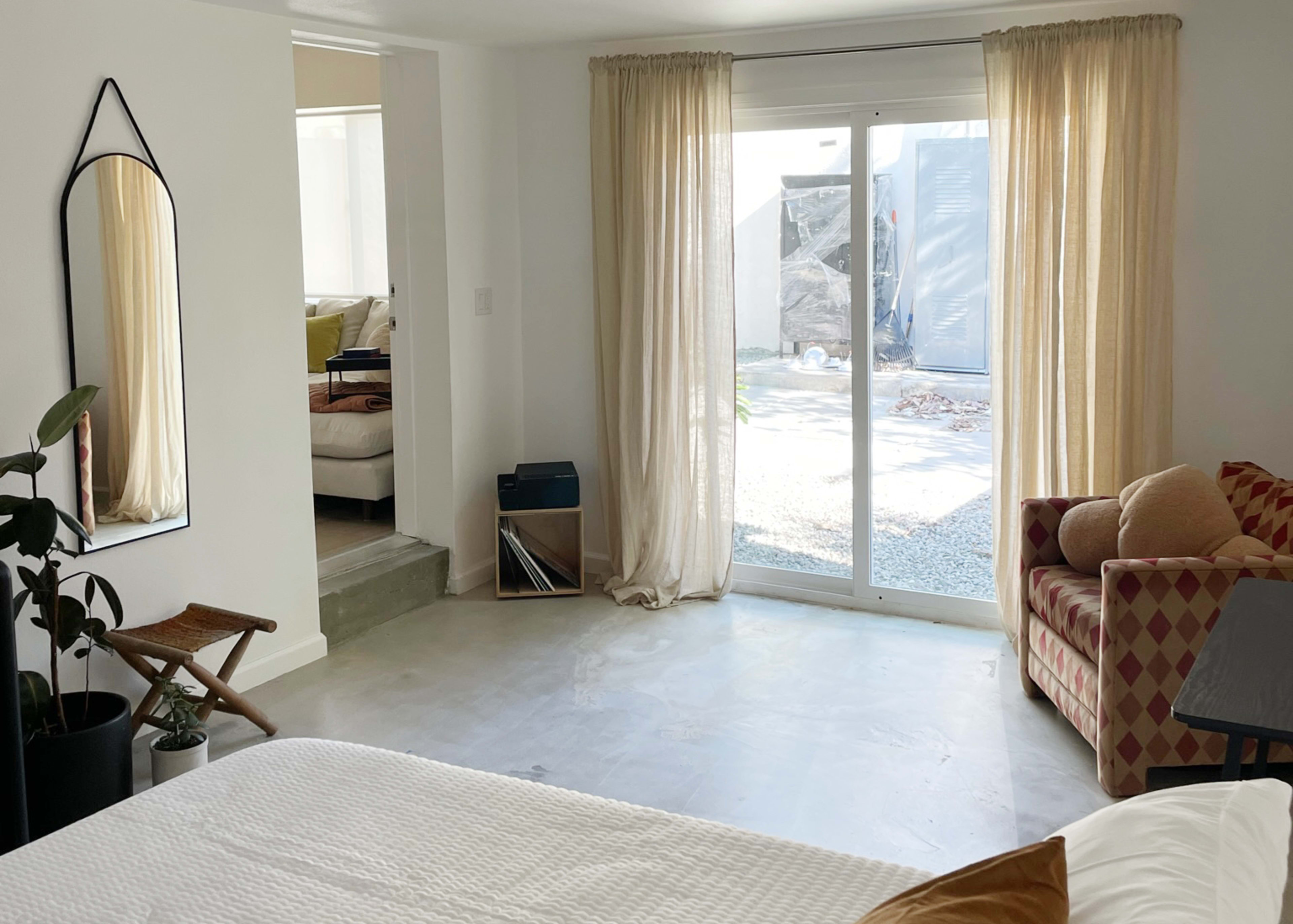 A sunlit bedroom features a bed, a mirror on the wall, a couch, and sliding glass doors leading to a patio area.
