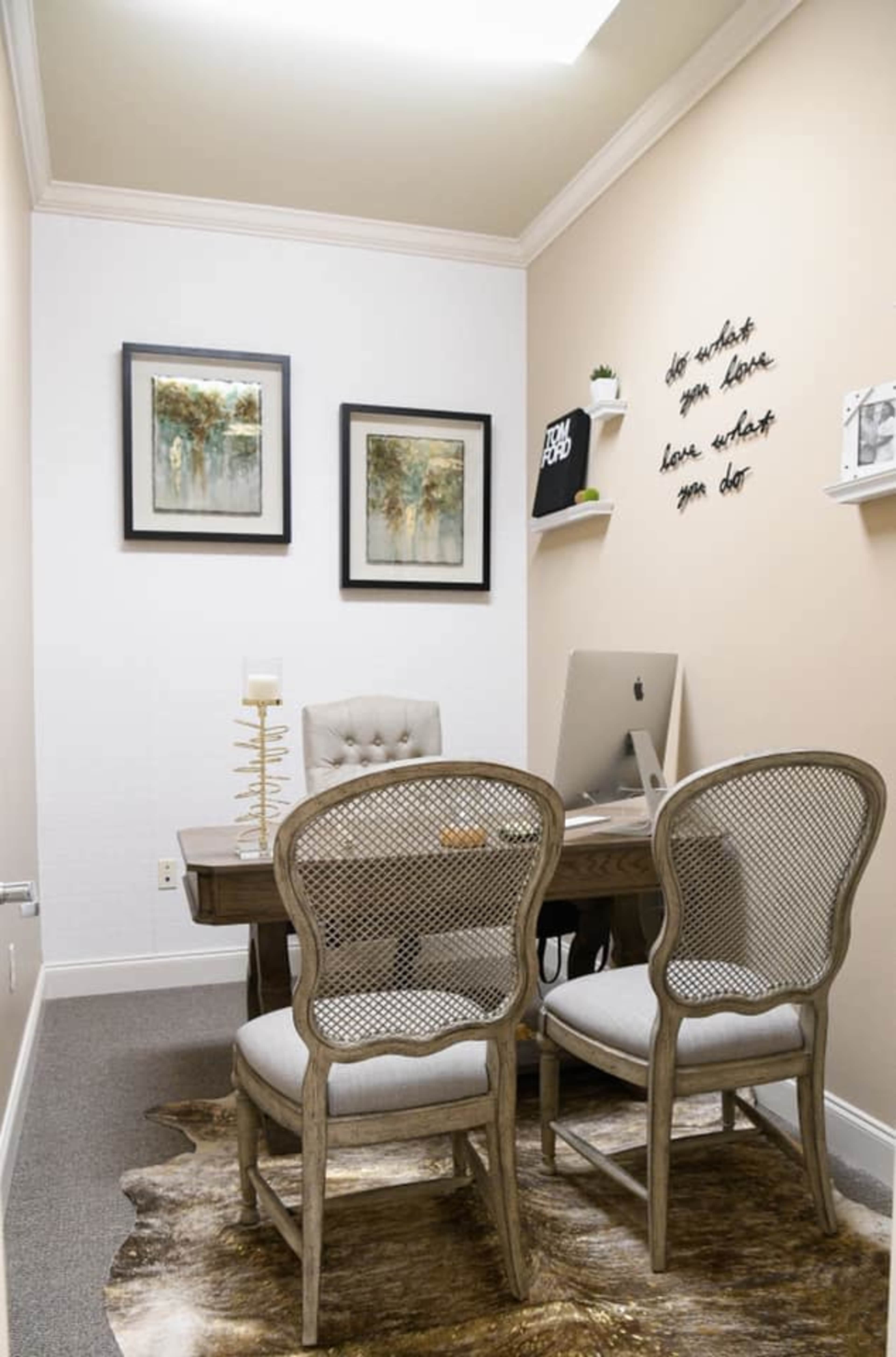 A small office space featuring a wooden desk with an Apple computer, two chairs, framed artworks on the walls, and a decorative wall quote.