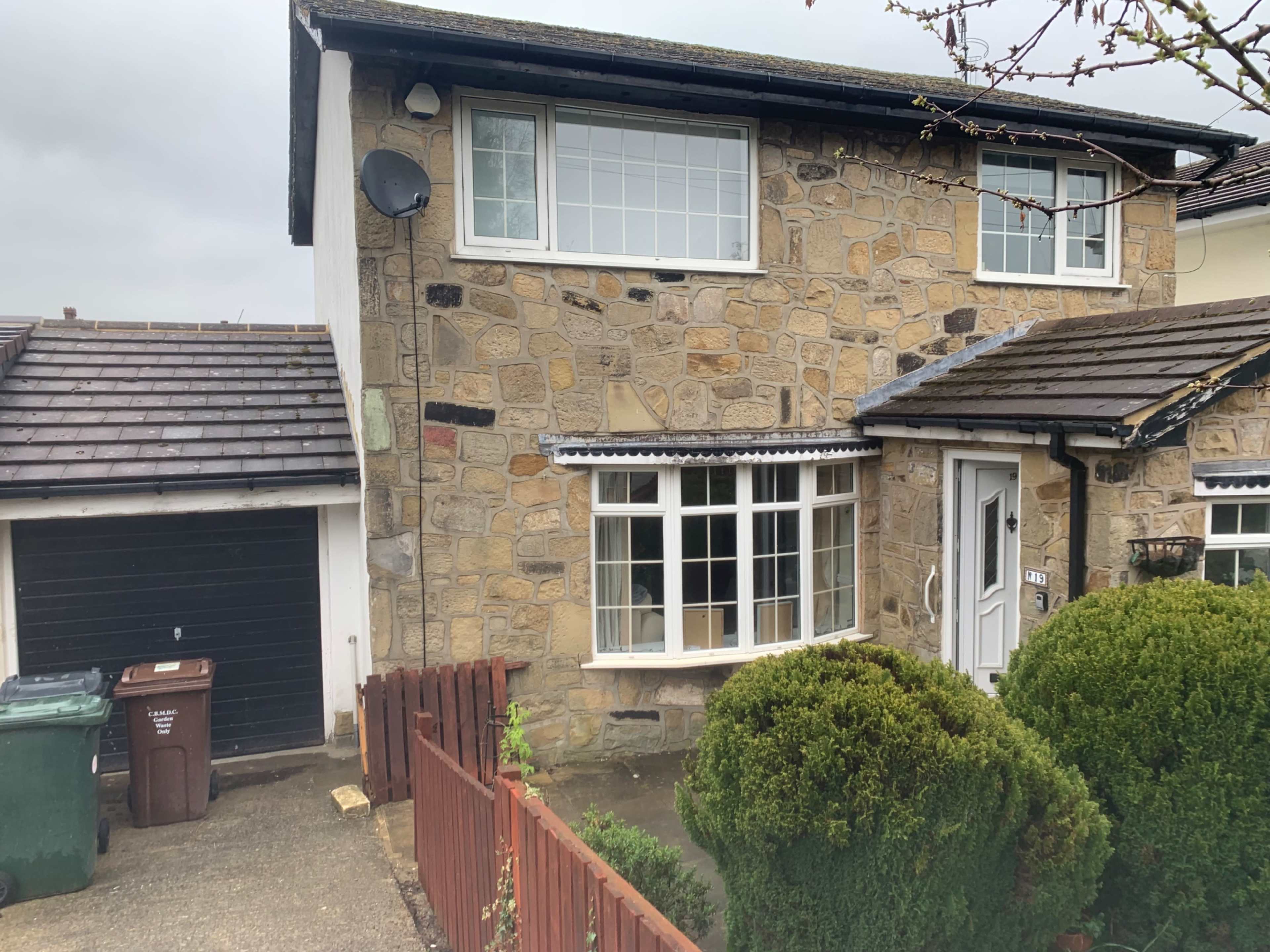 The image shows a two-story stone house with a front garden, a garage on the left, and a couple of green trash bins near the driveway.