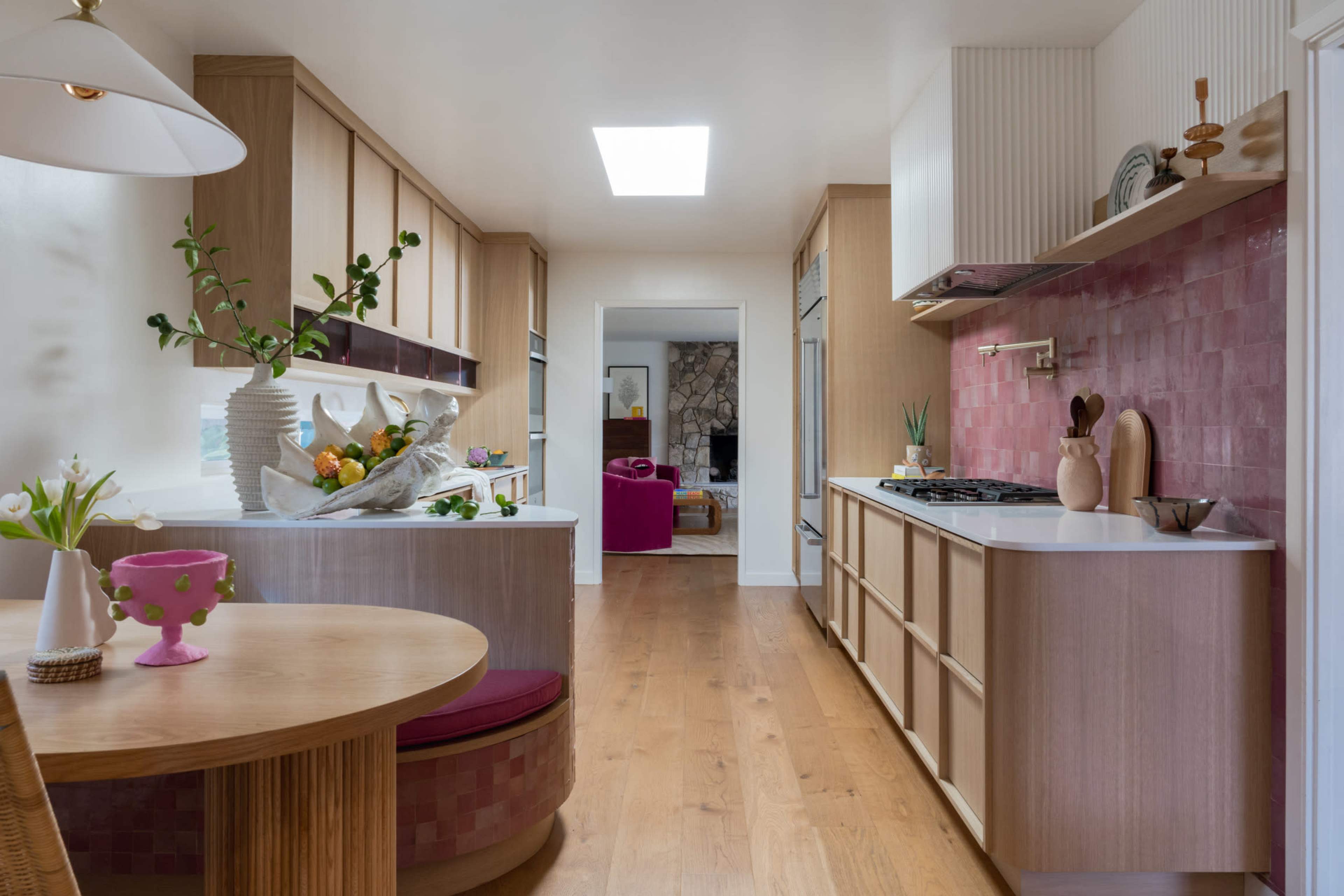 The image shows a modern kitchen with light wood cabinetry, a central round table, and a pink tiled backsplash.