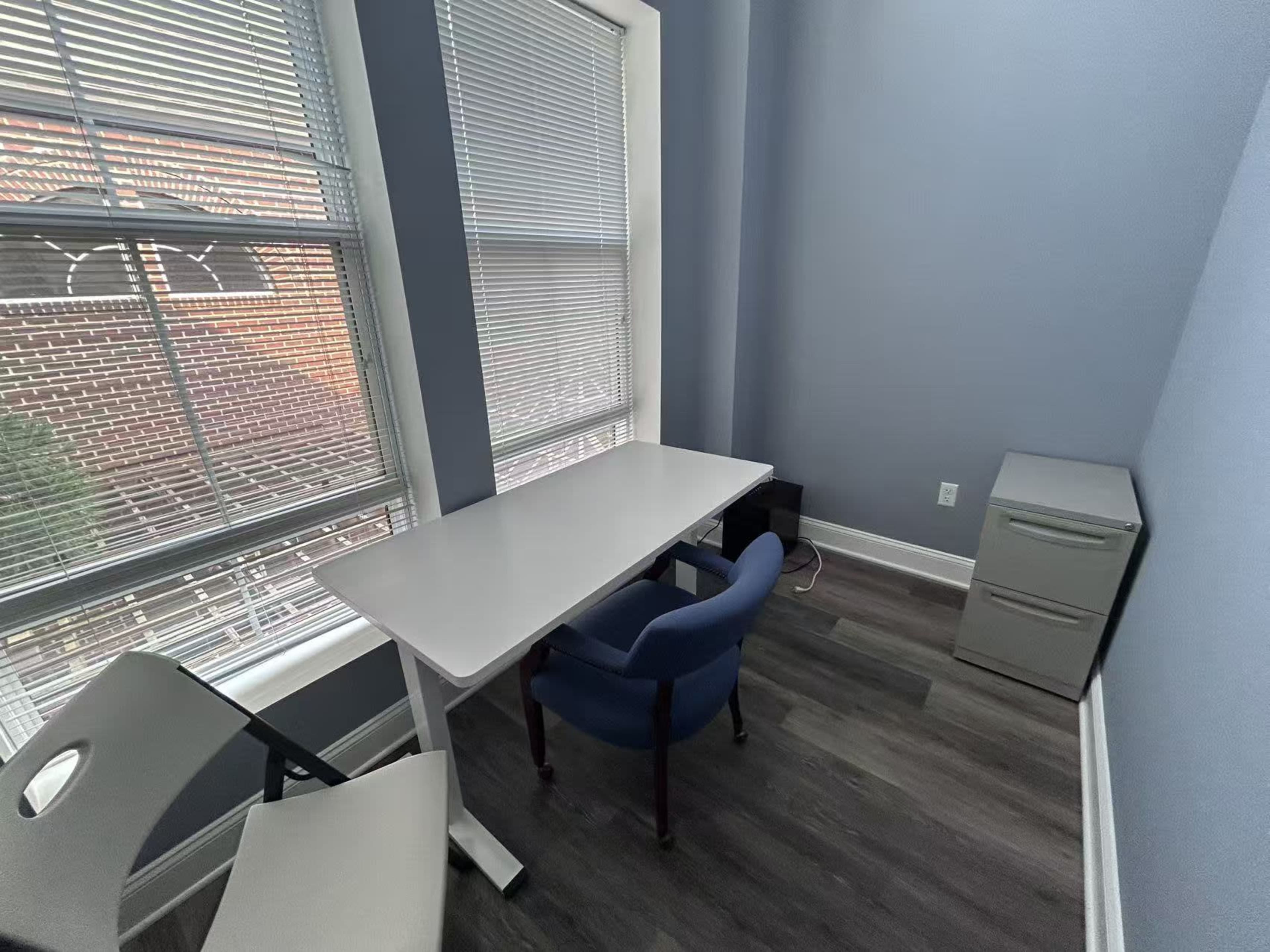 The image shows a small office space with a white table, a blue chair, and a gray filing cabinet, illuminated by natural light from two large windows.