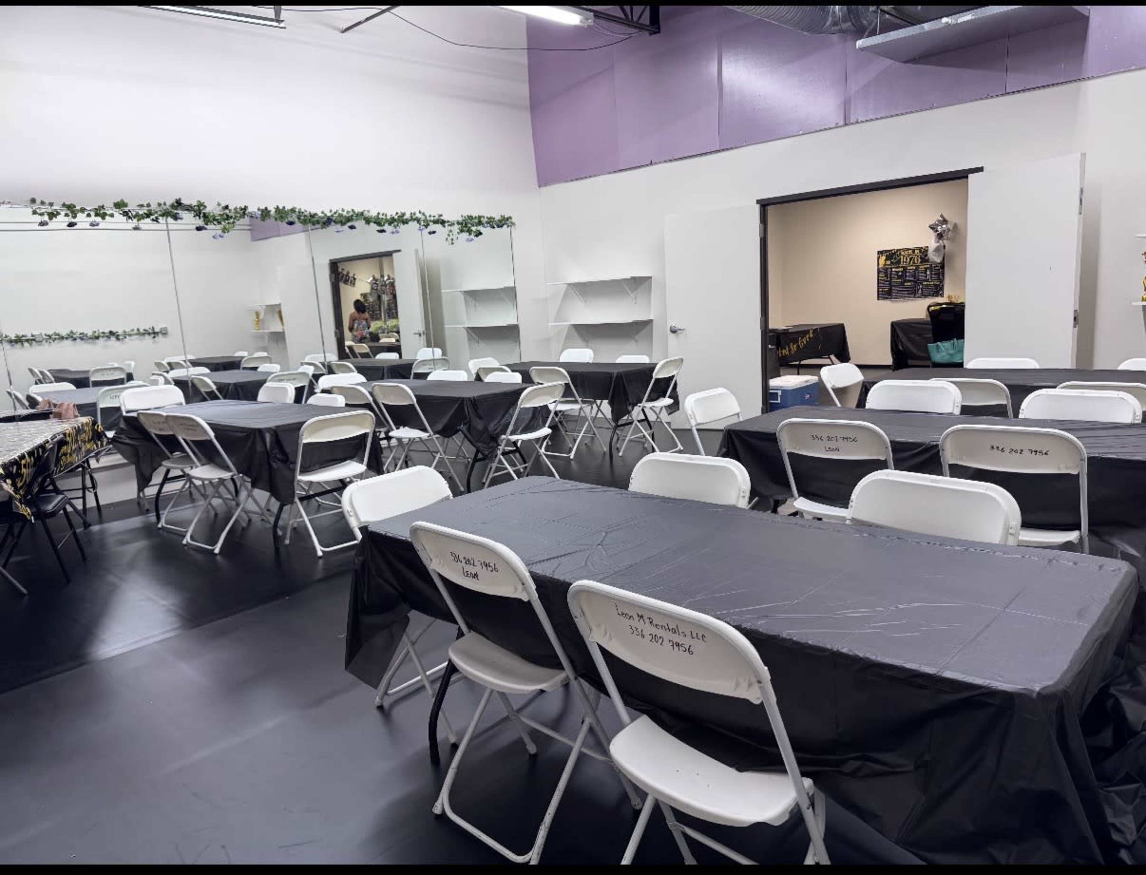 The image shows a classroom or event space with several rows of tables covered in black tablecloths and white folding chairs around them, along with a mirror on one wall and an entrance to another room in the background.