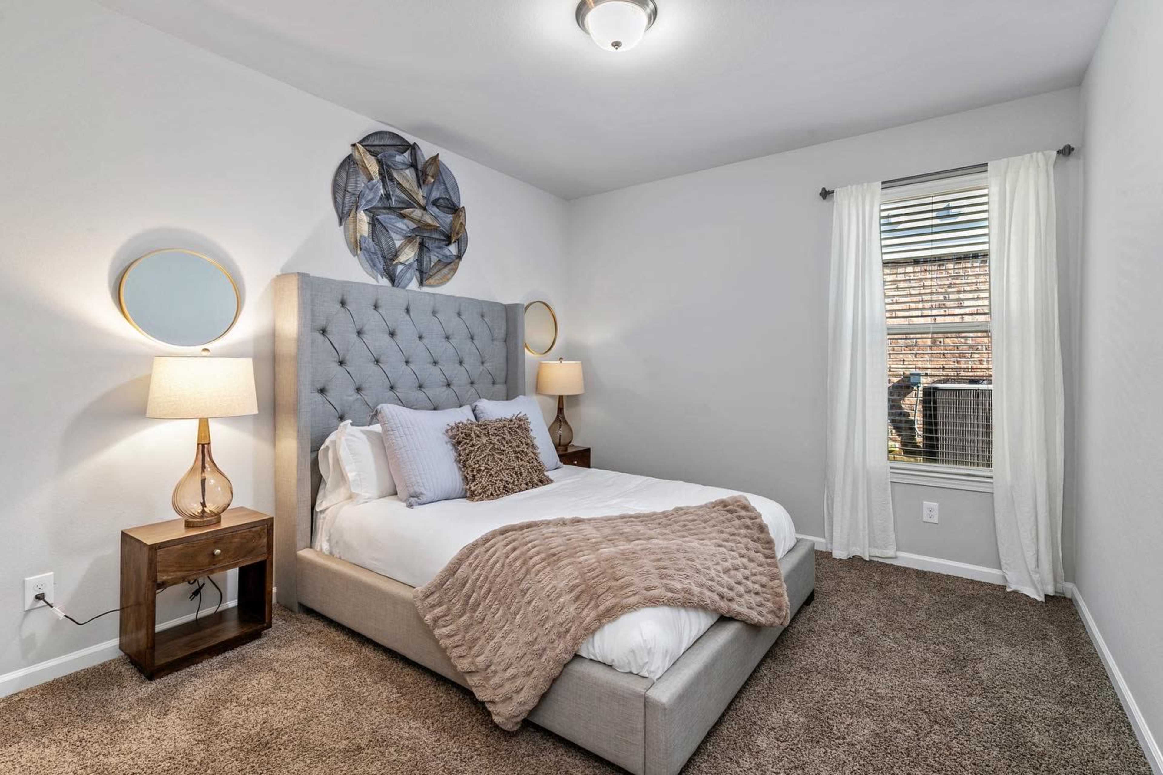 A neatly arranged bedroom featuring a tufted gray bed frame, two bedside tables with lamps, a decorative wall piece, and a window with sheer curtains.