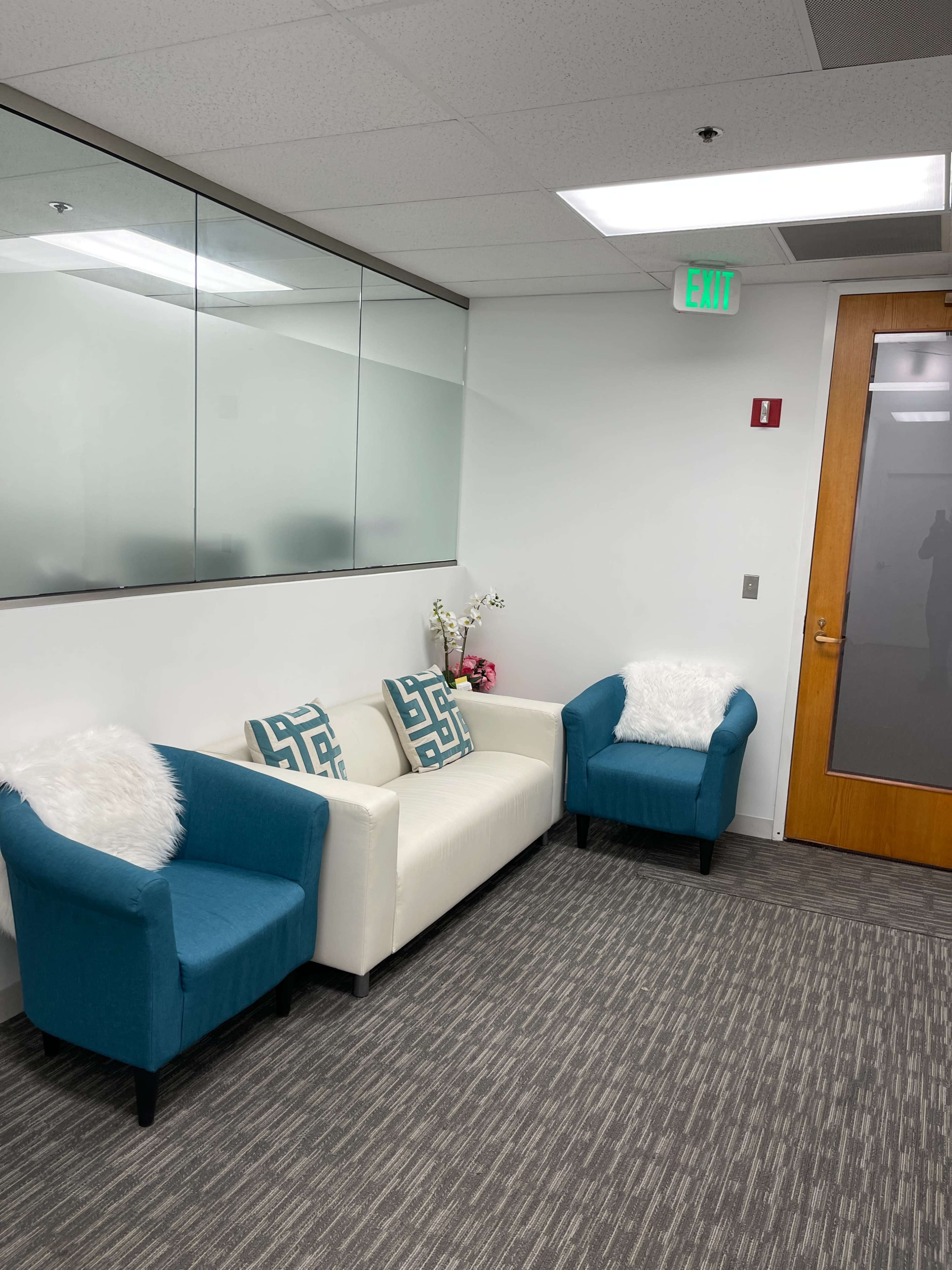 The image shows a modern waiting area with a white sofa and two turquoise chairs, adorned with decorative pillows and a small plant on a table.