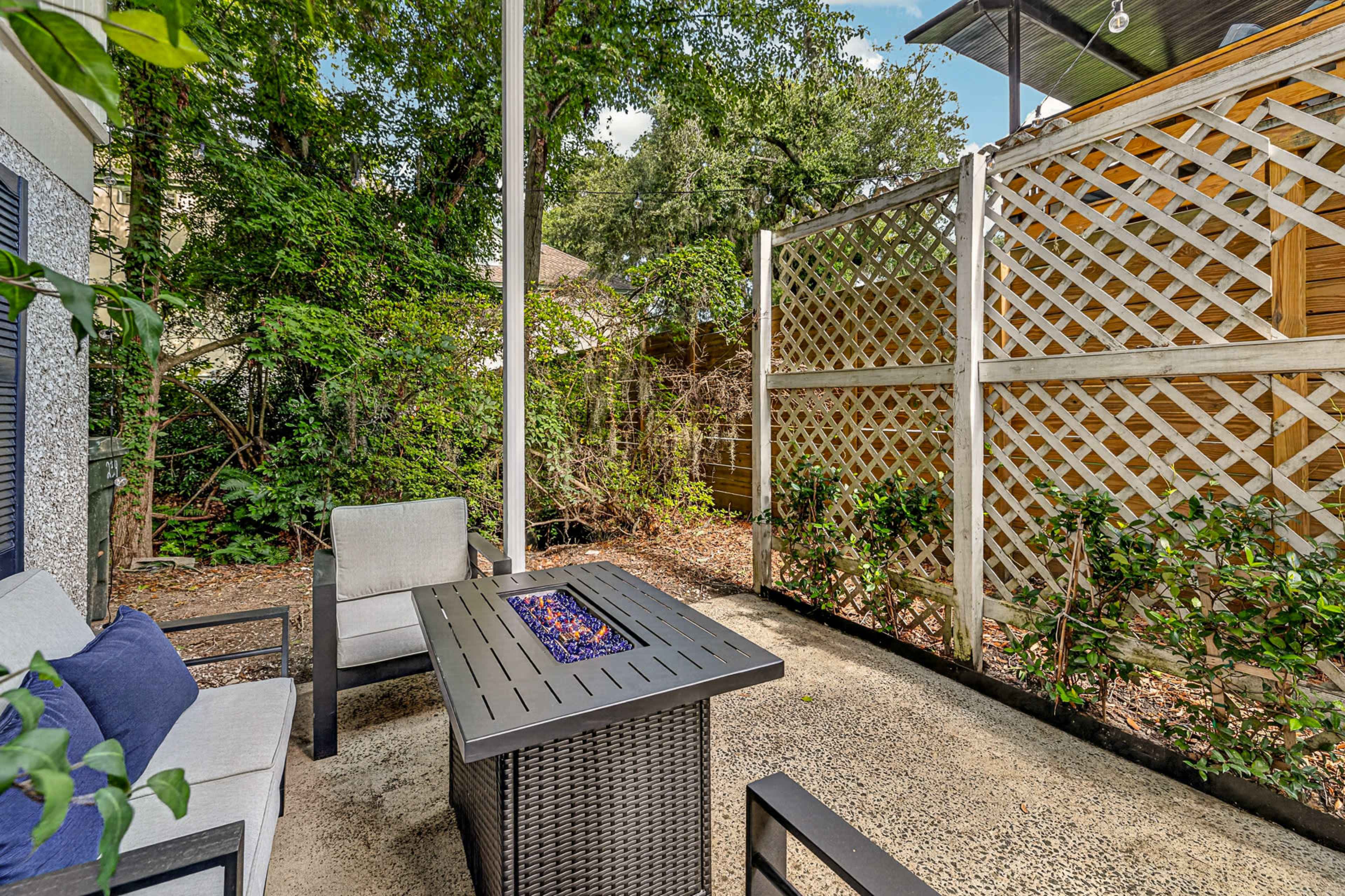 The image shows a small outdoor seating area with two chairs and a table featuring a fire element, surrounded by green foliage and a wooden lattice fence.
