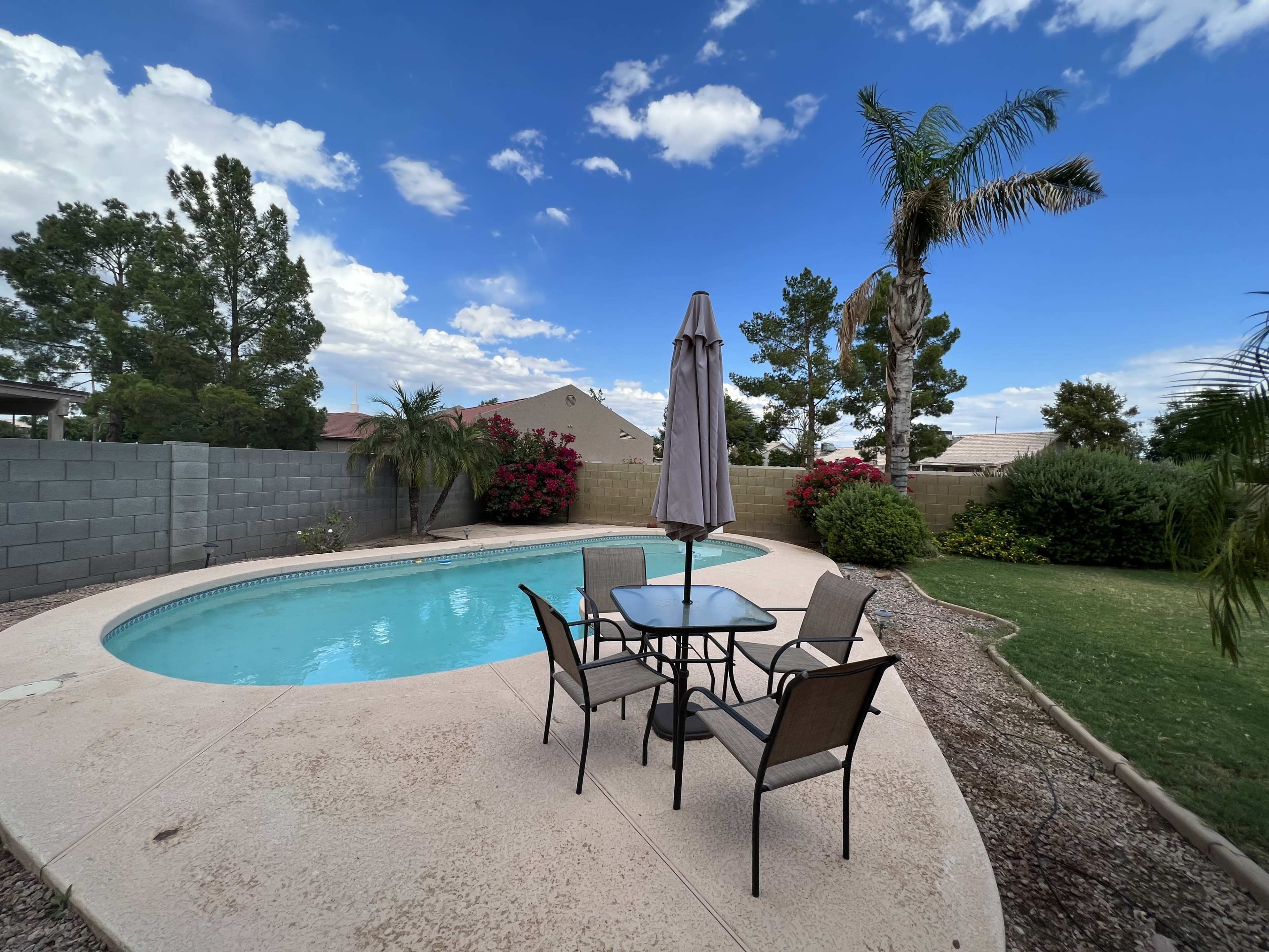 A patio area with a table and chairs is situated beside a swimming pool, surrounded by greenery and flowering bushes under a partly cloudy sky.