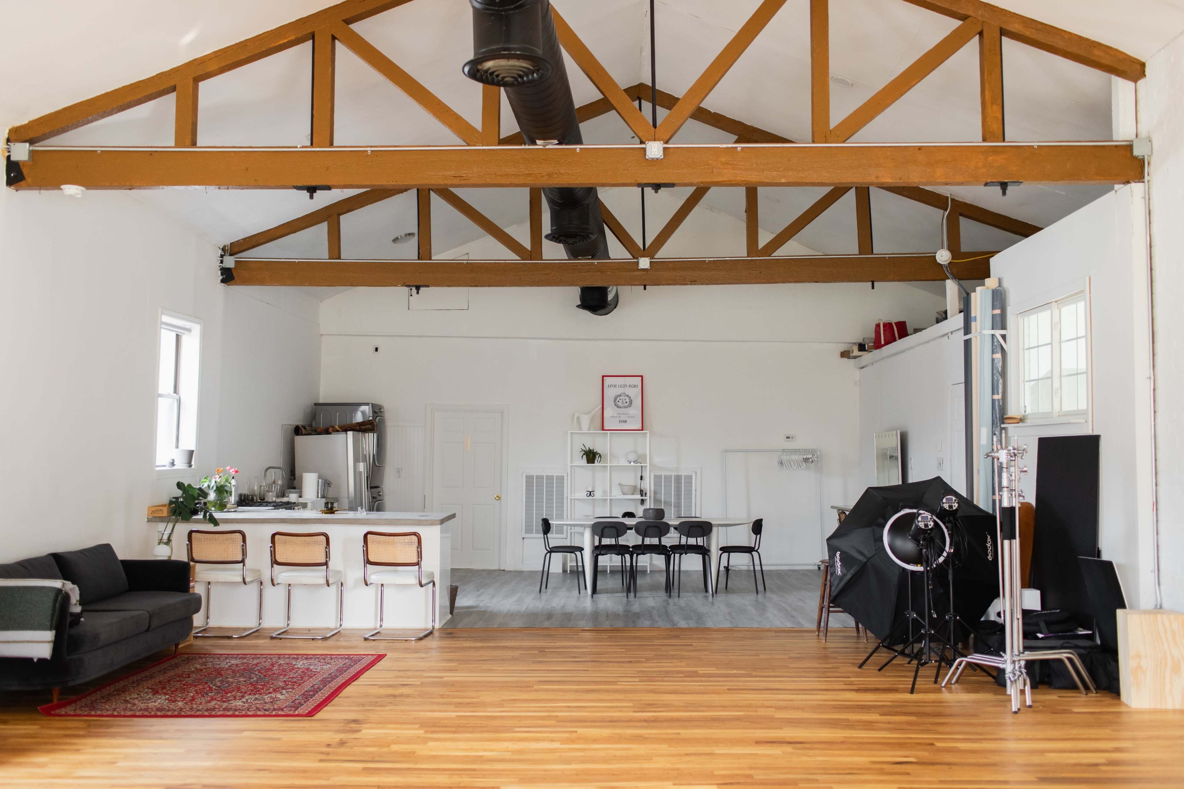 A spacious, well-lit interior with wooden beams, featuring a seating area with a couch, a kitchen counter, a dining table, and photography equipment in the corner.
