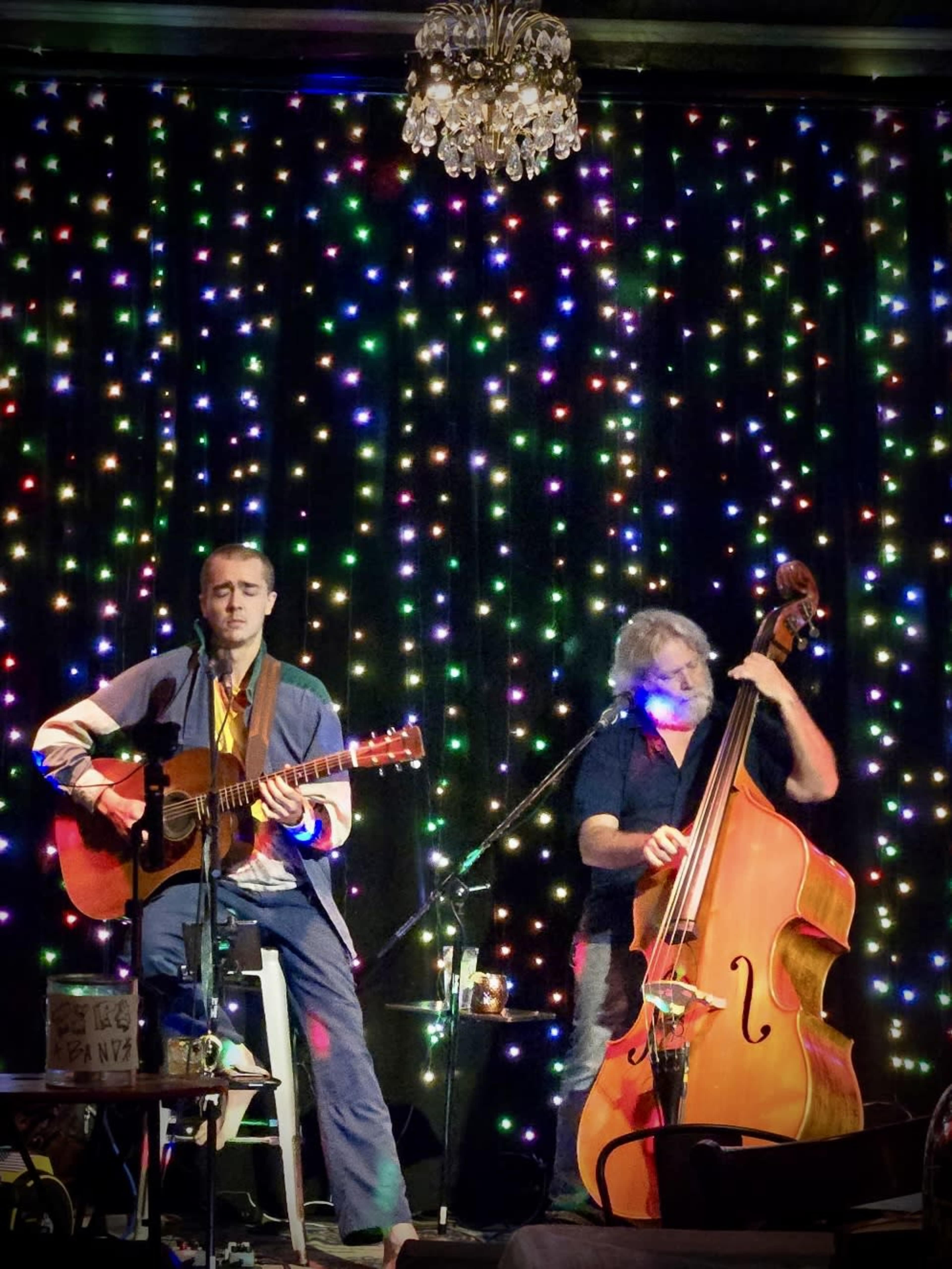 A musician plays an acoustic guitar while sitting on a stool, accompanied by a bassist, in front of a backdrop of colorful lights.