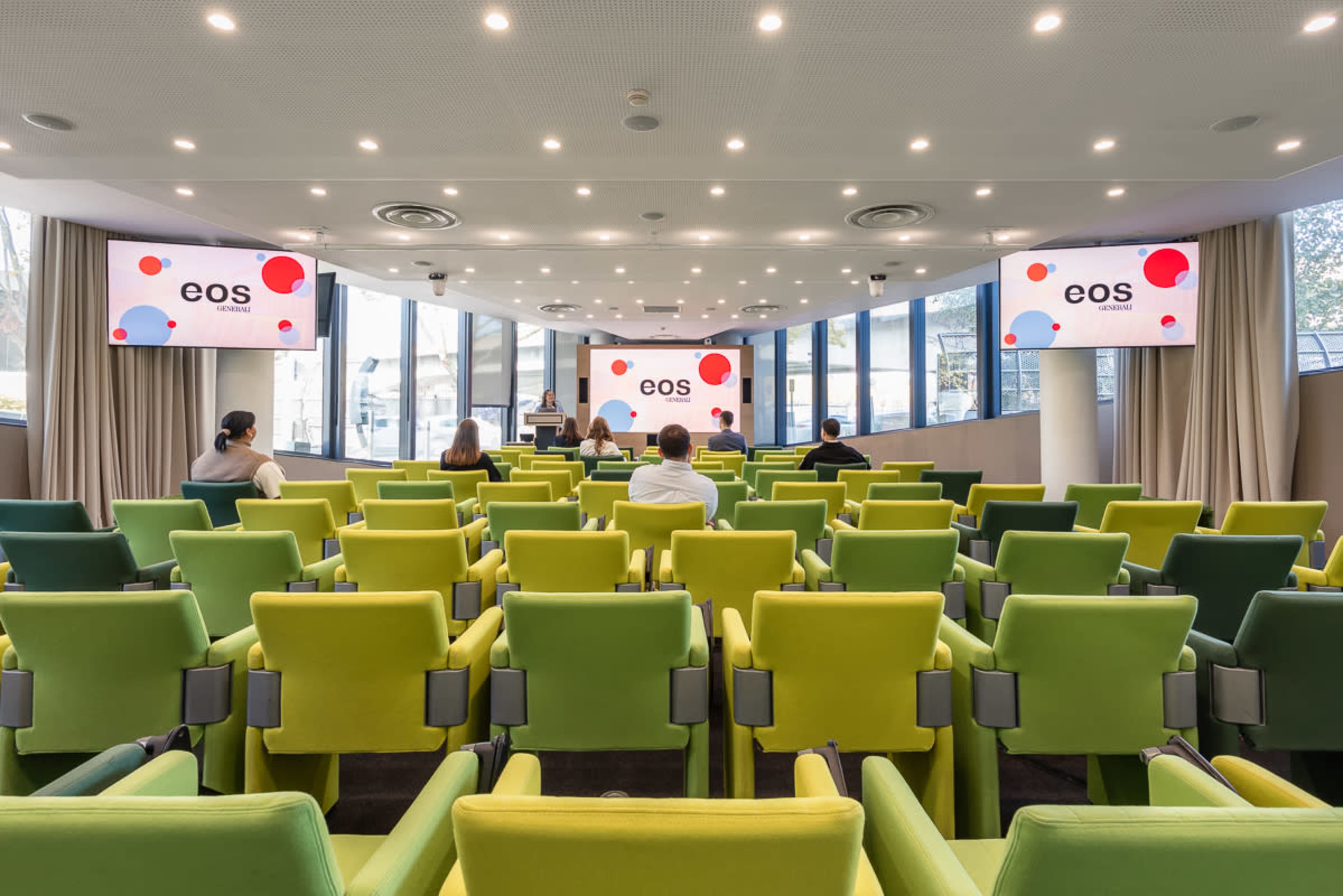 The image shows a modern conference room with a series of green and yellow chairs arranged in rows facing a stage with large screens displaying the logo "eos."