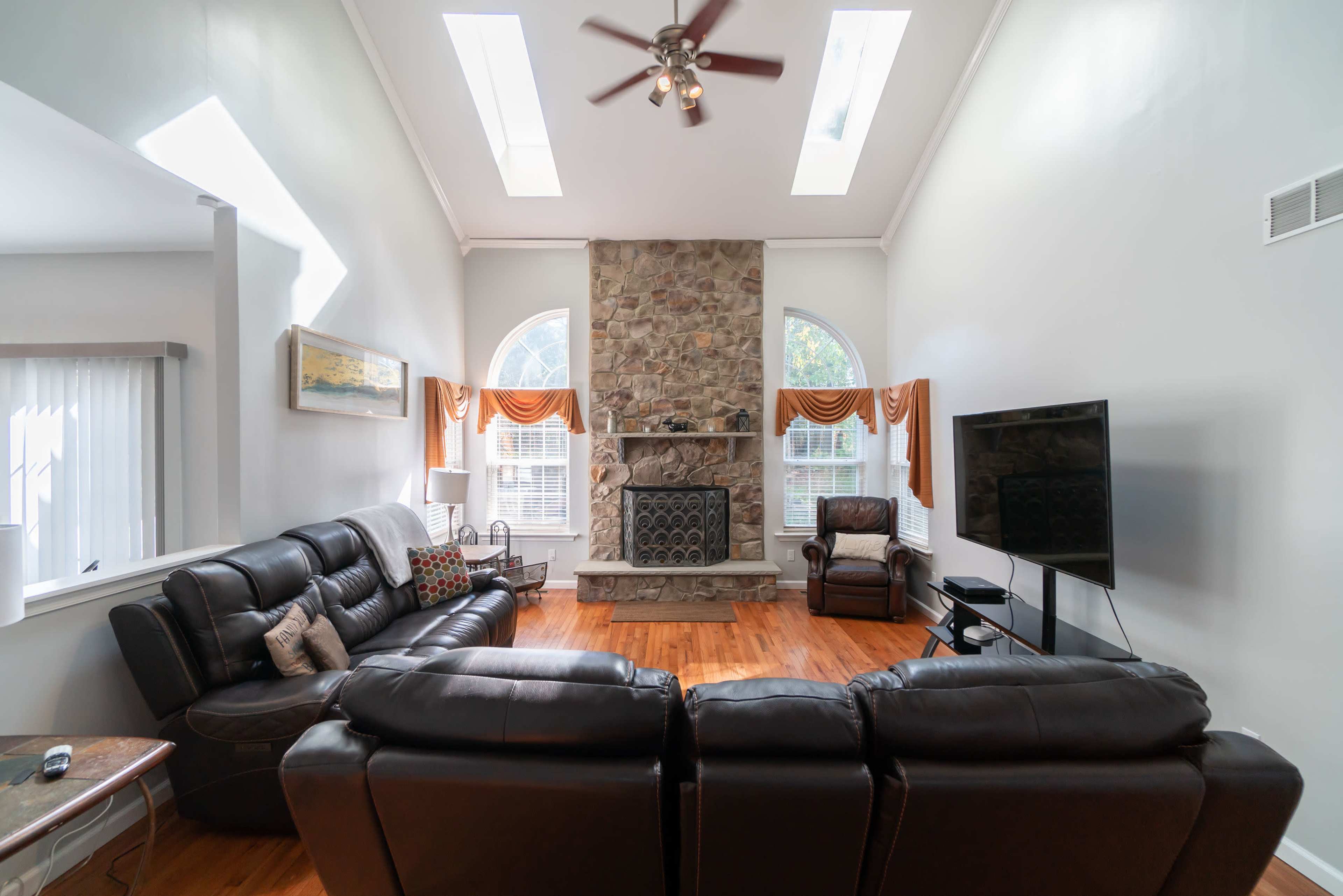A spacious living room features a stone fireplace, large windows, and leather furniture arranged around a central coffee table.