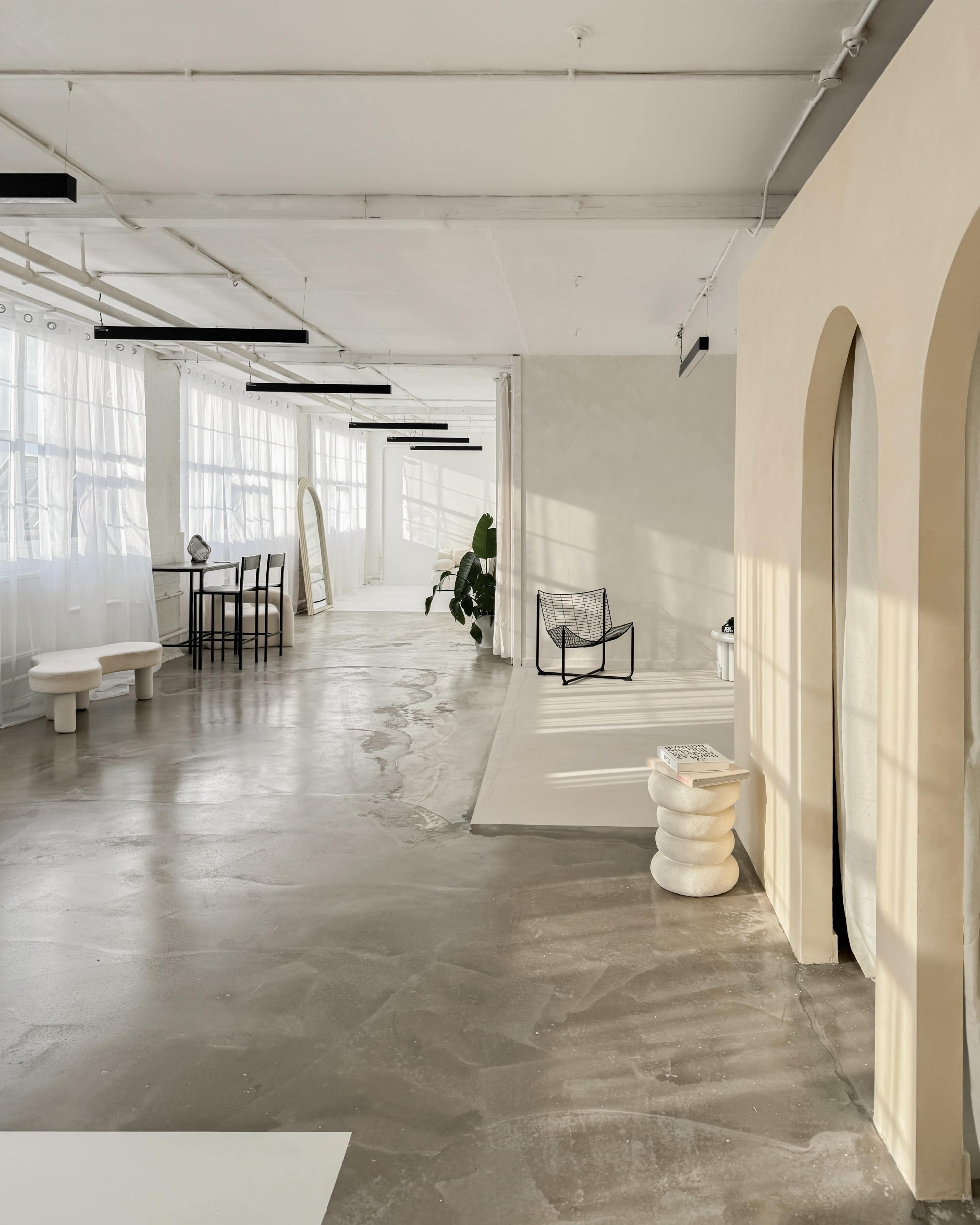 The image shows a spacious, well-lit interior with minimalist furniture, large windows, and a polished concrete floor.