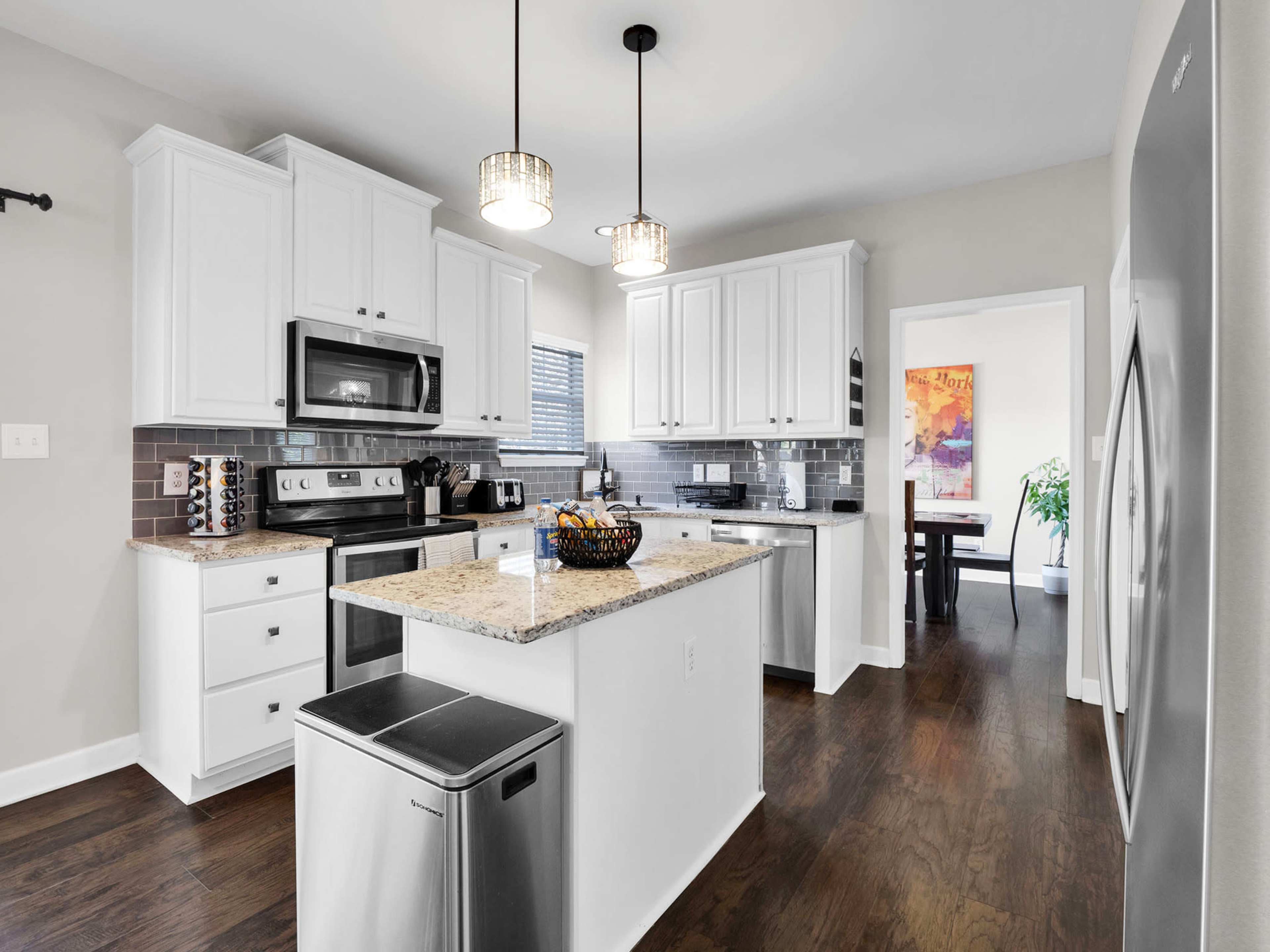 A modern kitchen featuring white cabinetry, a granite island with a fruit bowl, stainless steel appliances, and dark hardwood flooring.