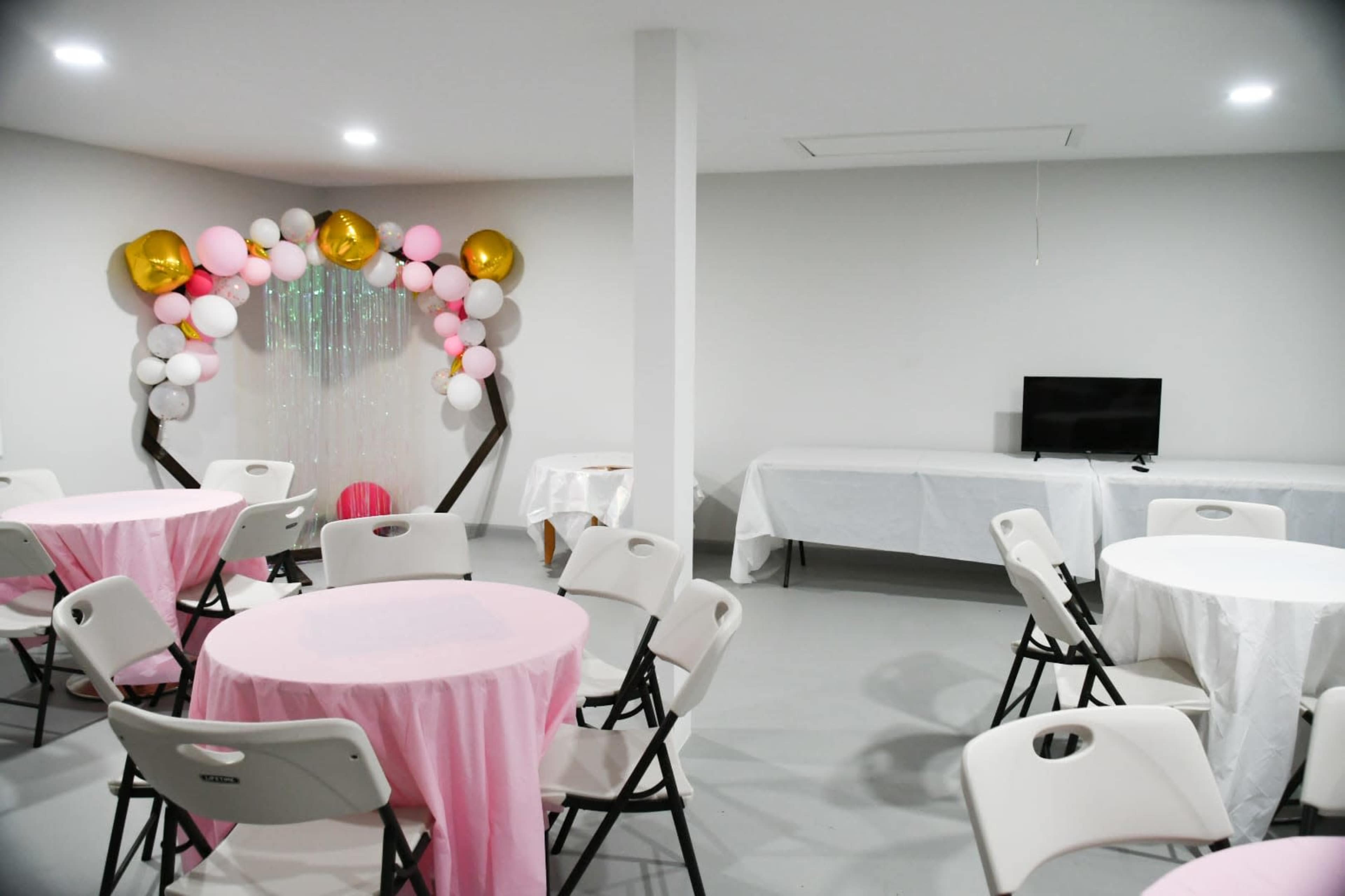 A decorated party room features round tables covered in pink cloth, with an arch of balloons and a television in the background.