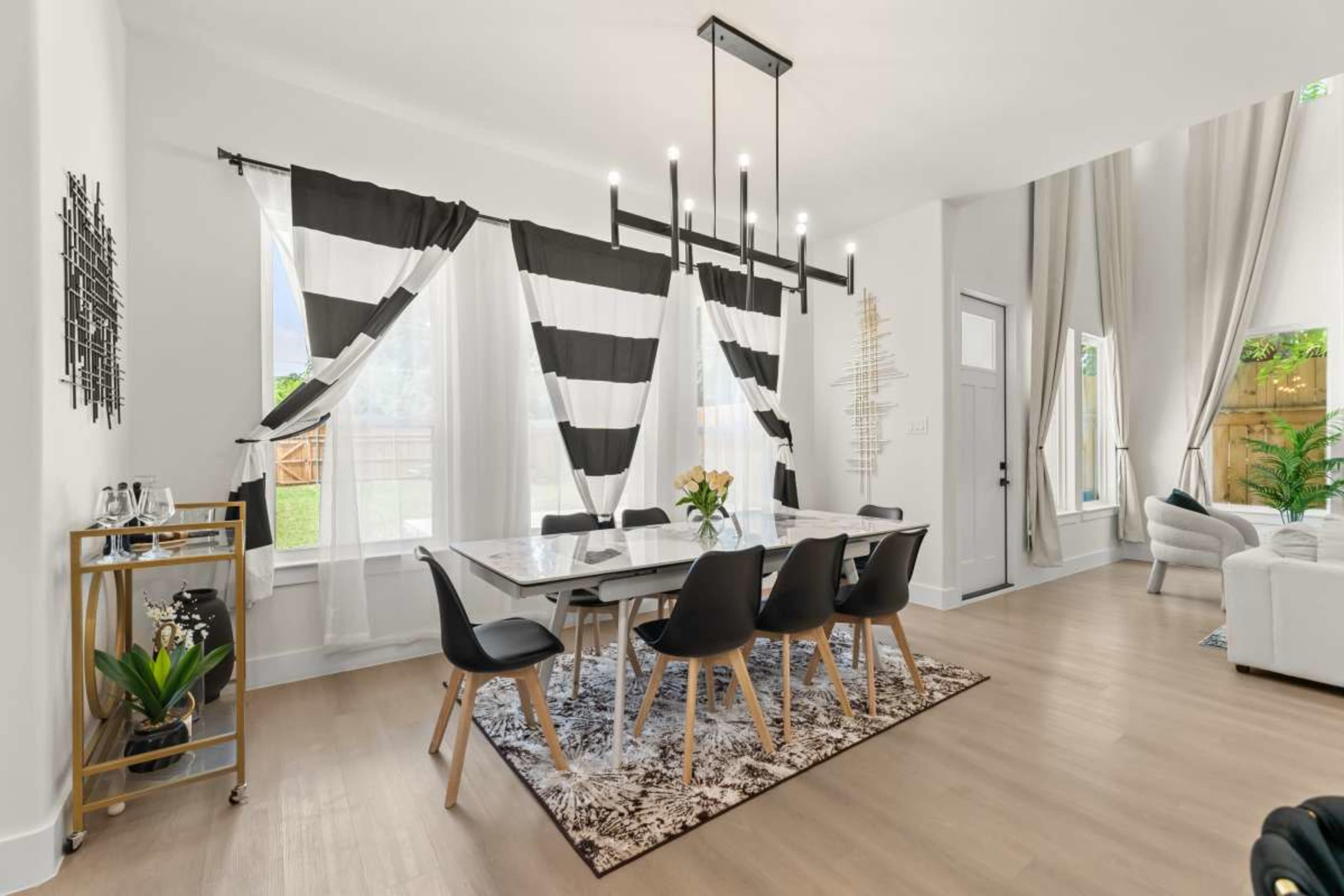The image shows a modern dining area featuring a long table surrounded by black chairs, with large striped curtains and a decorative rug.