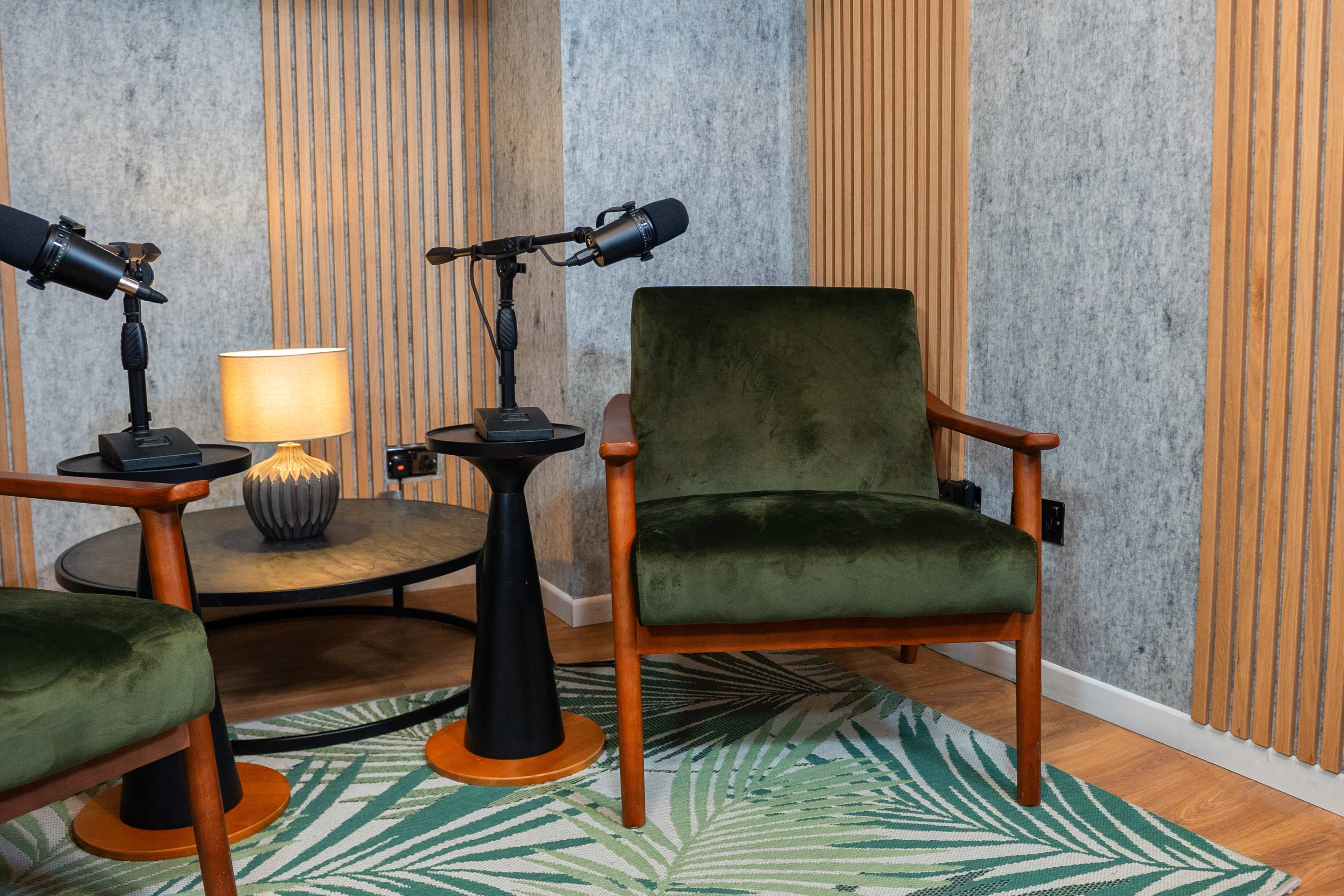 A small seating area featuring two green upholstered chairs, a round black side table with a lamp, and microphones on stands in a room with textured walls and wood accents.