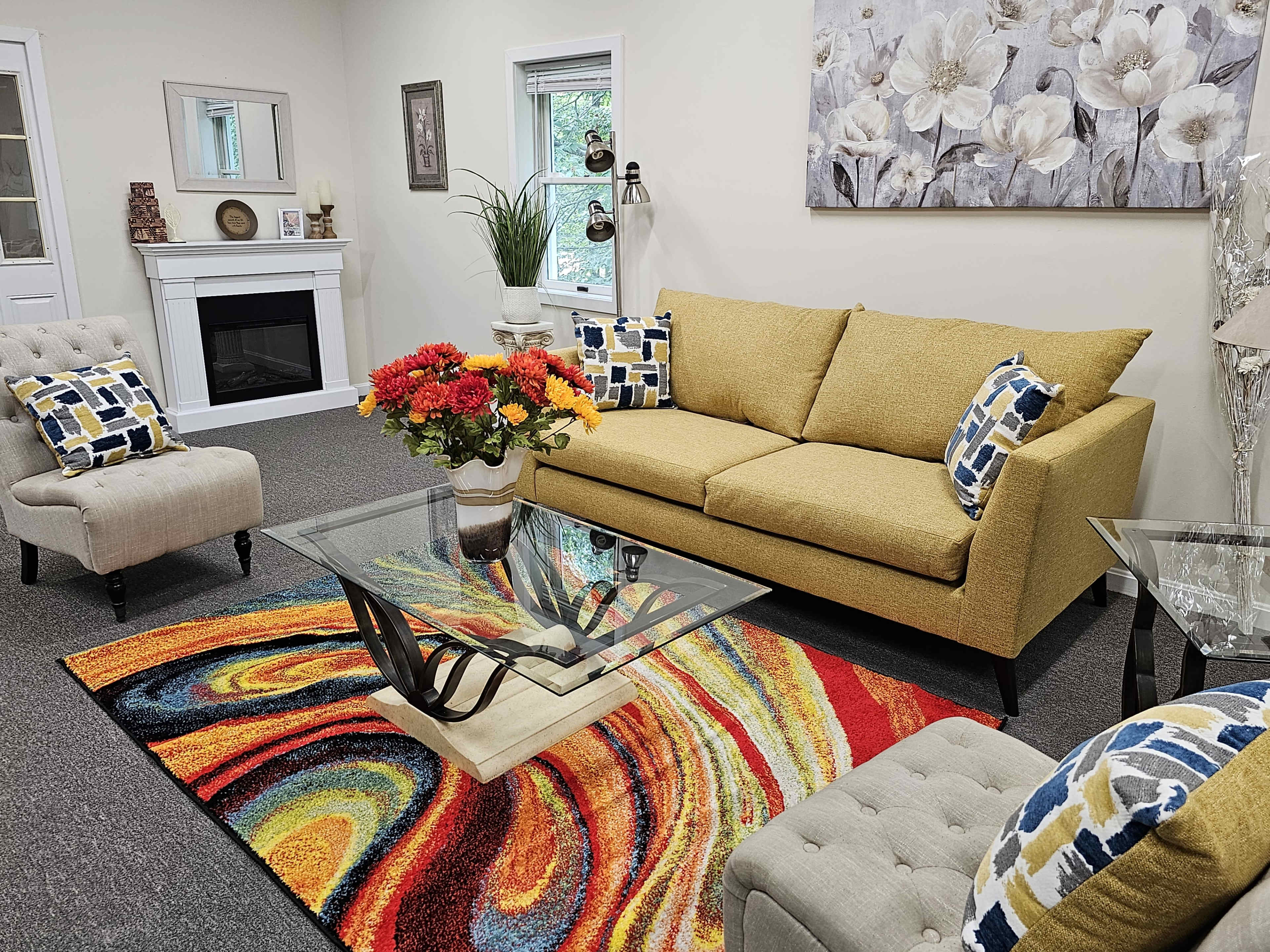 The image shows a cozy living room with a yellow sofa, two accent chairs, a glass coffee table, and a colorful swirling rug.